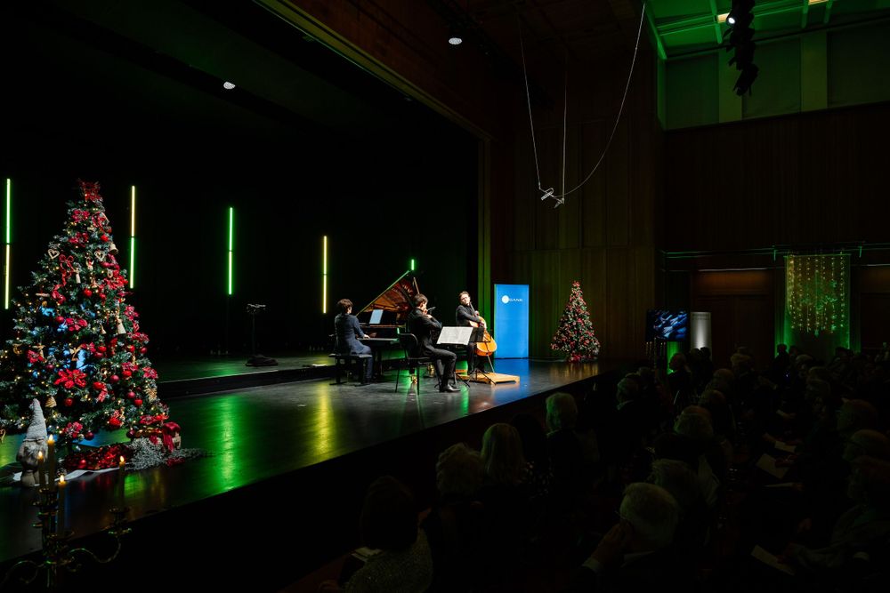 A dimly lit concert hall with Christmas trees and green lighting features musicians performing on stage.