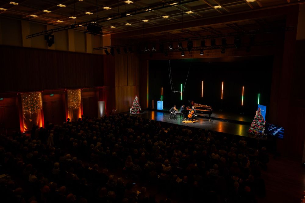 A concert hall filled with audience members watching performers on stage with decorative holiday lighting.