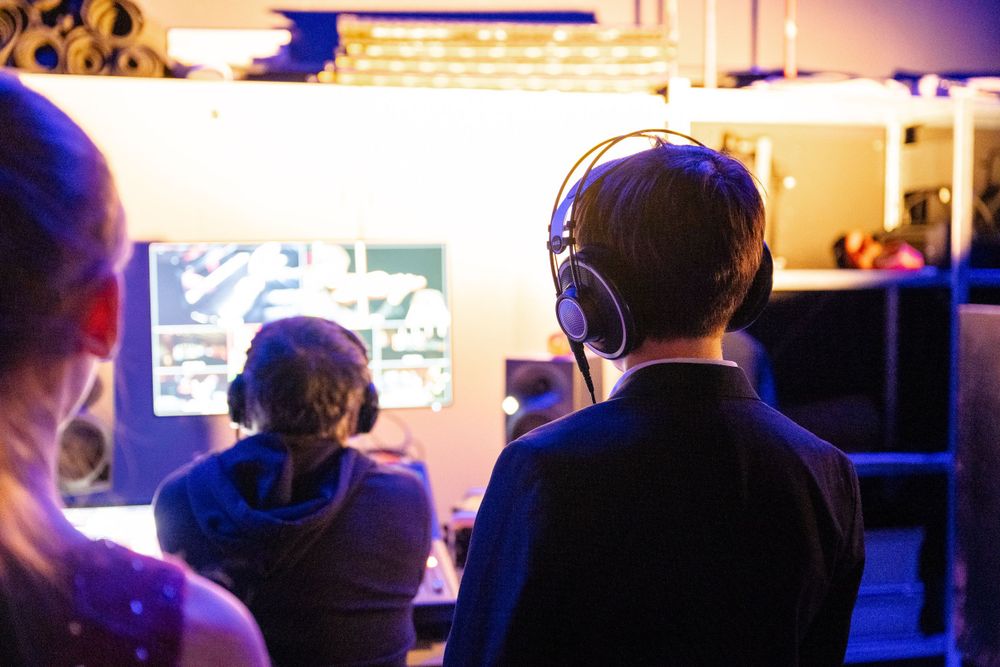 Production crew members watch monitors while wearing headsets in a dimly lit control room.