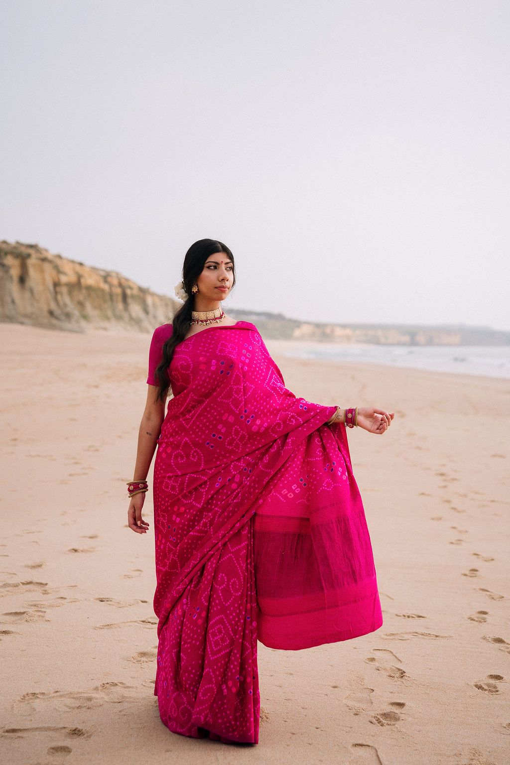 Person in bright pink traditional sari twirling on a beach with cliffs in the background.