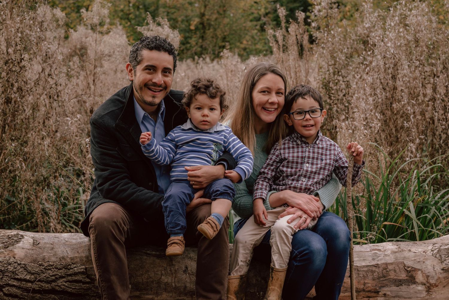 A family sits together outdoors on a fallen log surrounded by tall brown grass during golden hour.