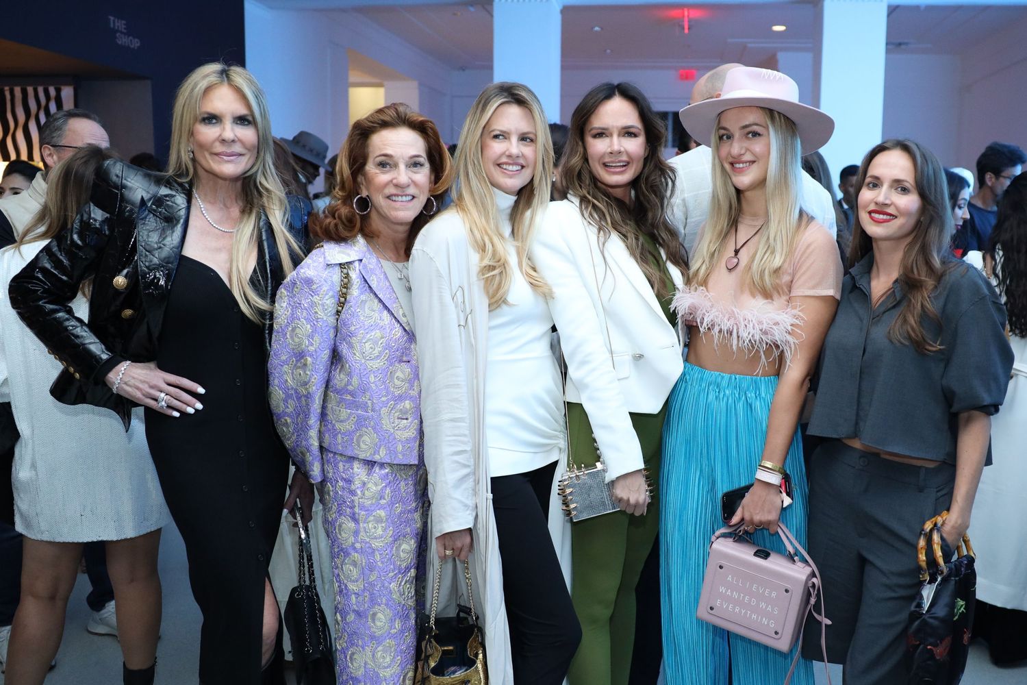 A group of stylishly dressed women pose together at an indoor social event wearing colorful dresses and accessories.