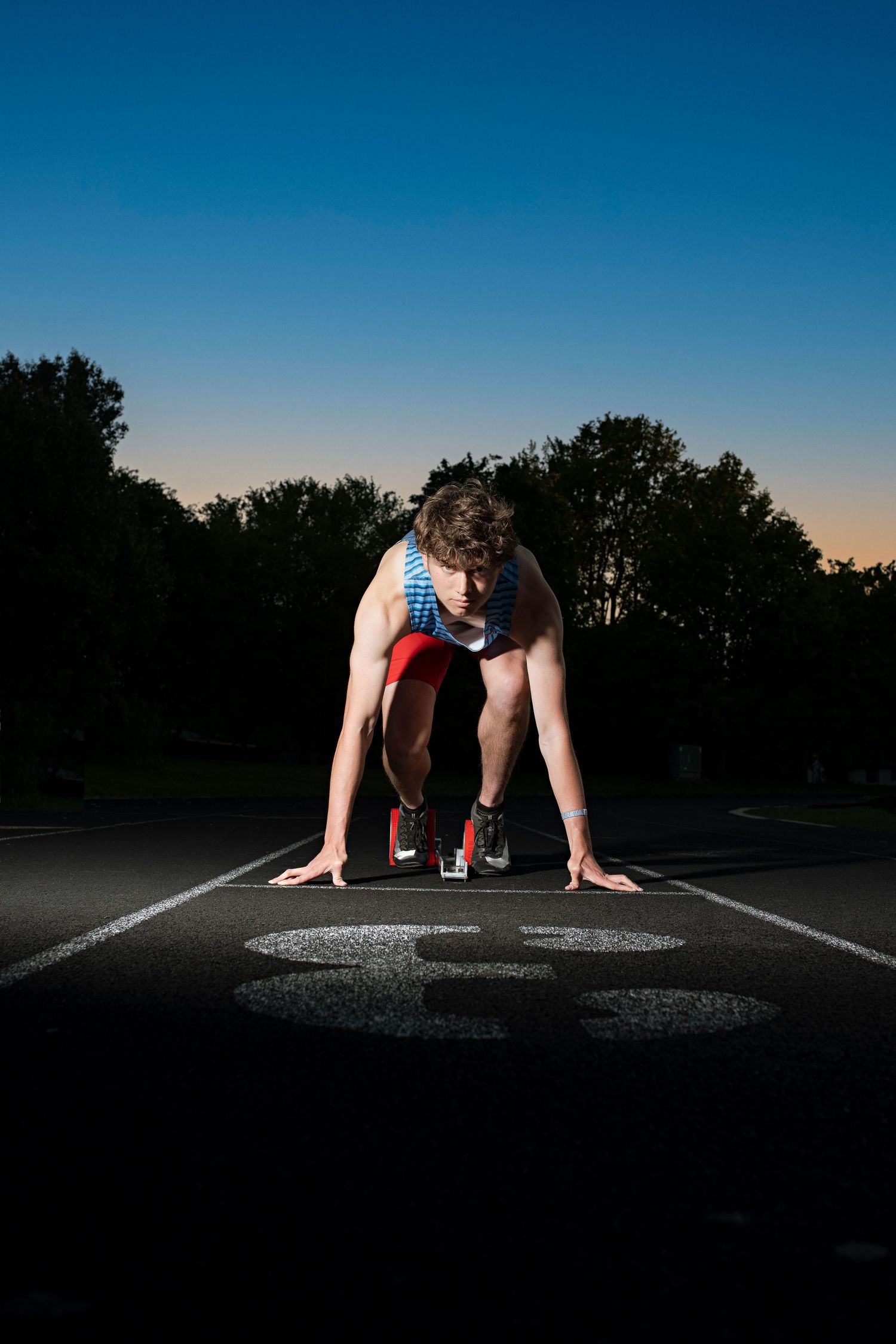 Athlete in starting position on a dark track at sunset with trees in the background.