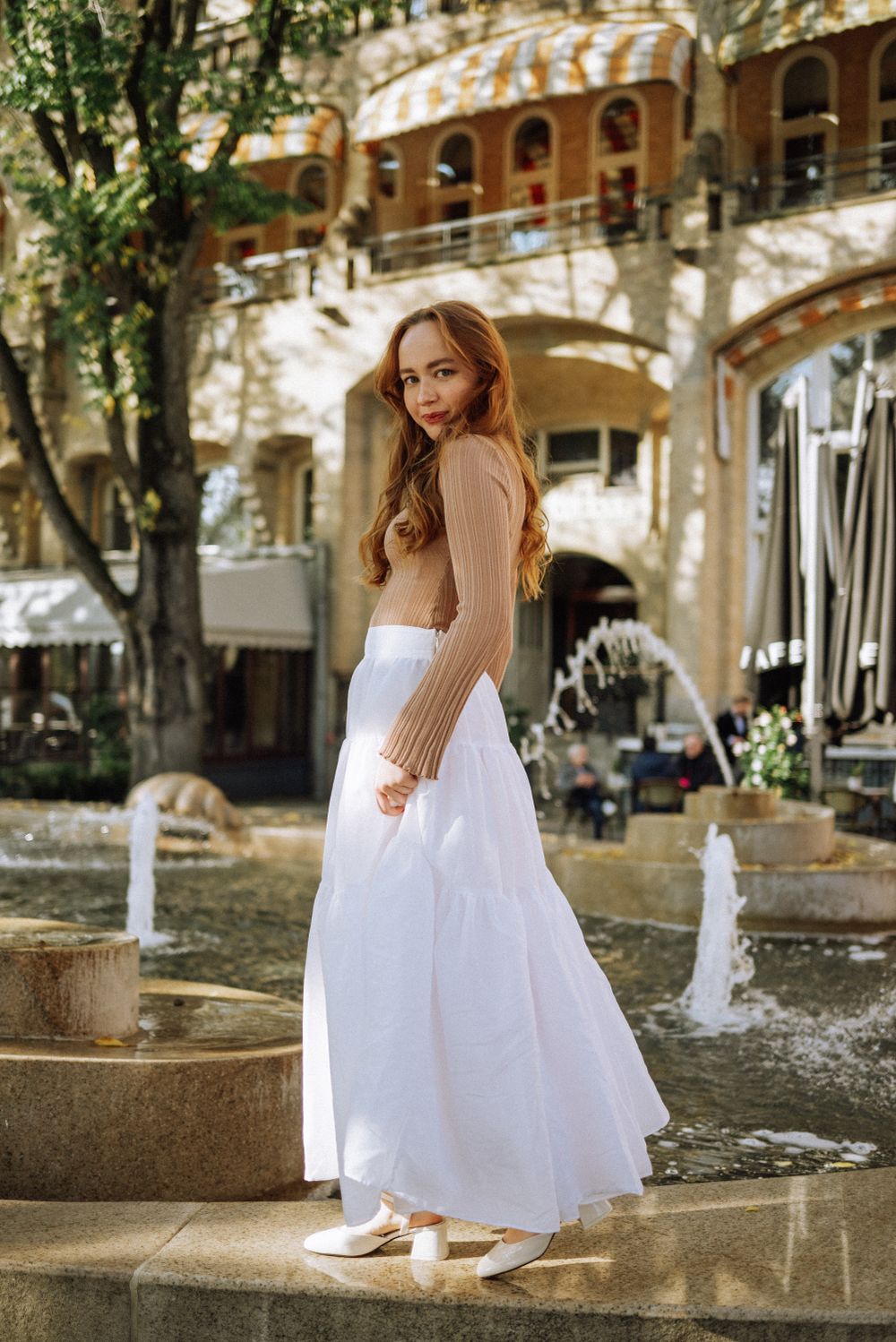 A person in a white maxi skirt and beige top stands by a fountain in a European courtyard.