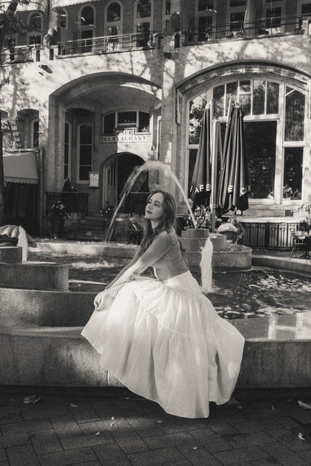 A black and white photo captures a dancer in a flowing white dress posing by a fountain.