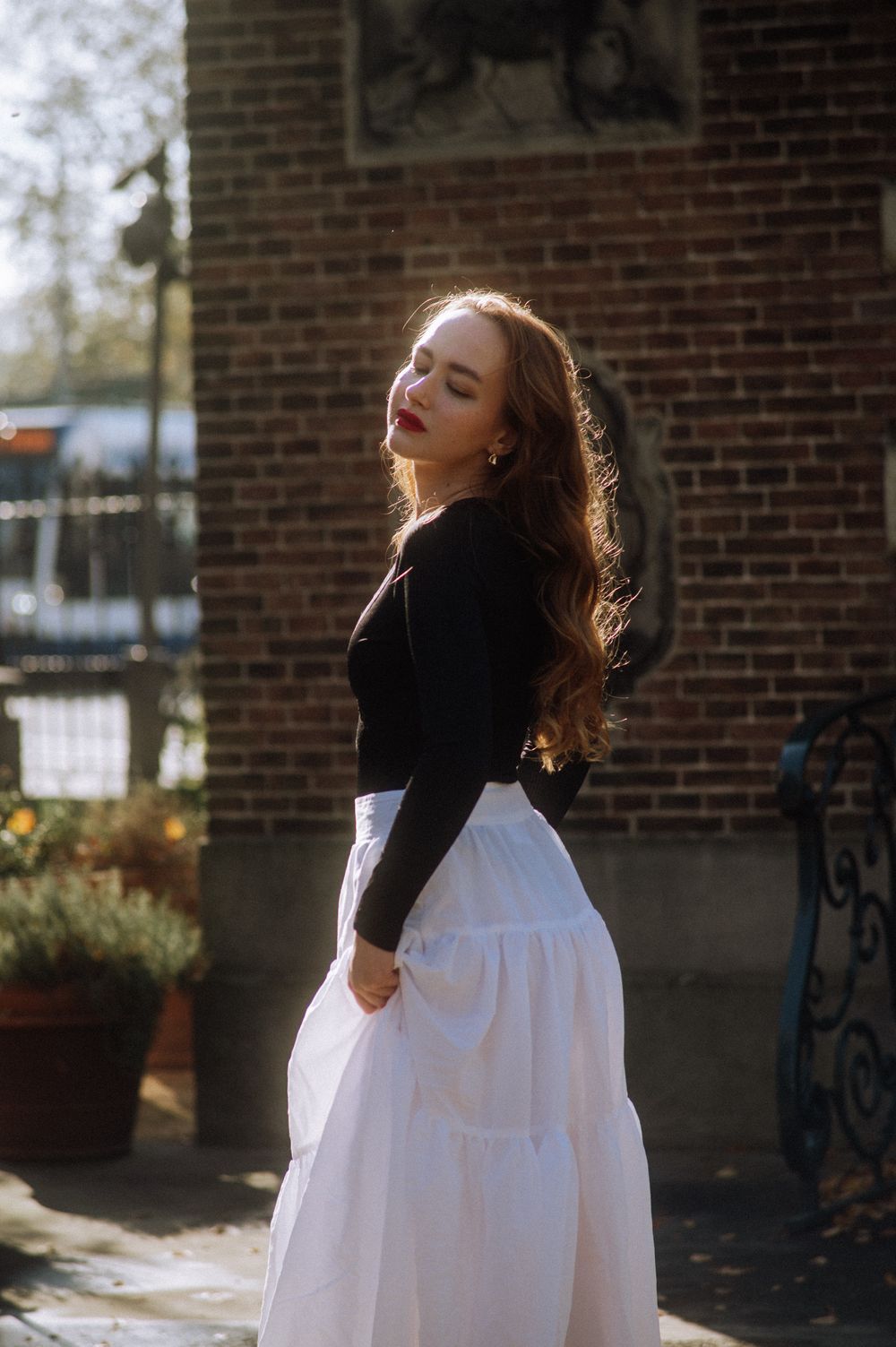 Sun-lit portrait of person in black top and white dress against brick building.