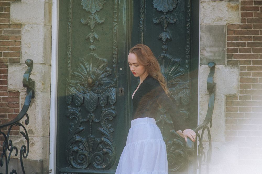 Person in white dress walking near ornate green doors and brick building.