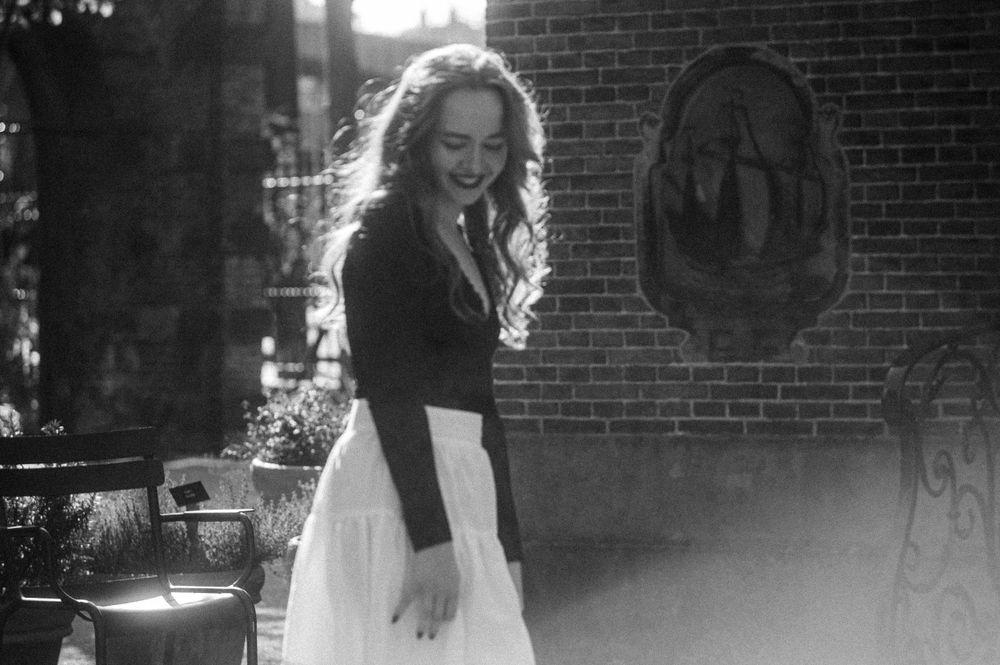 A woman in a black and white outfit poses dramatically in an outdoor setting with brick architecture.