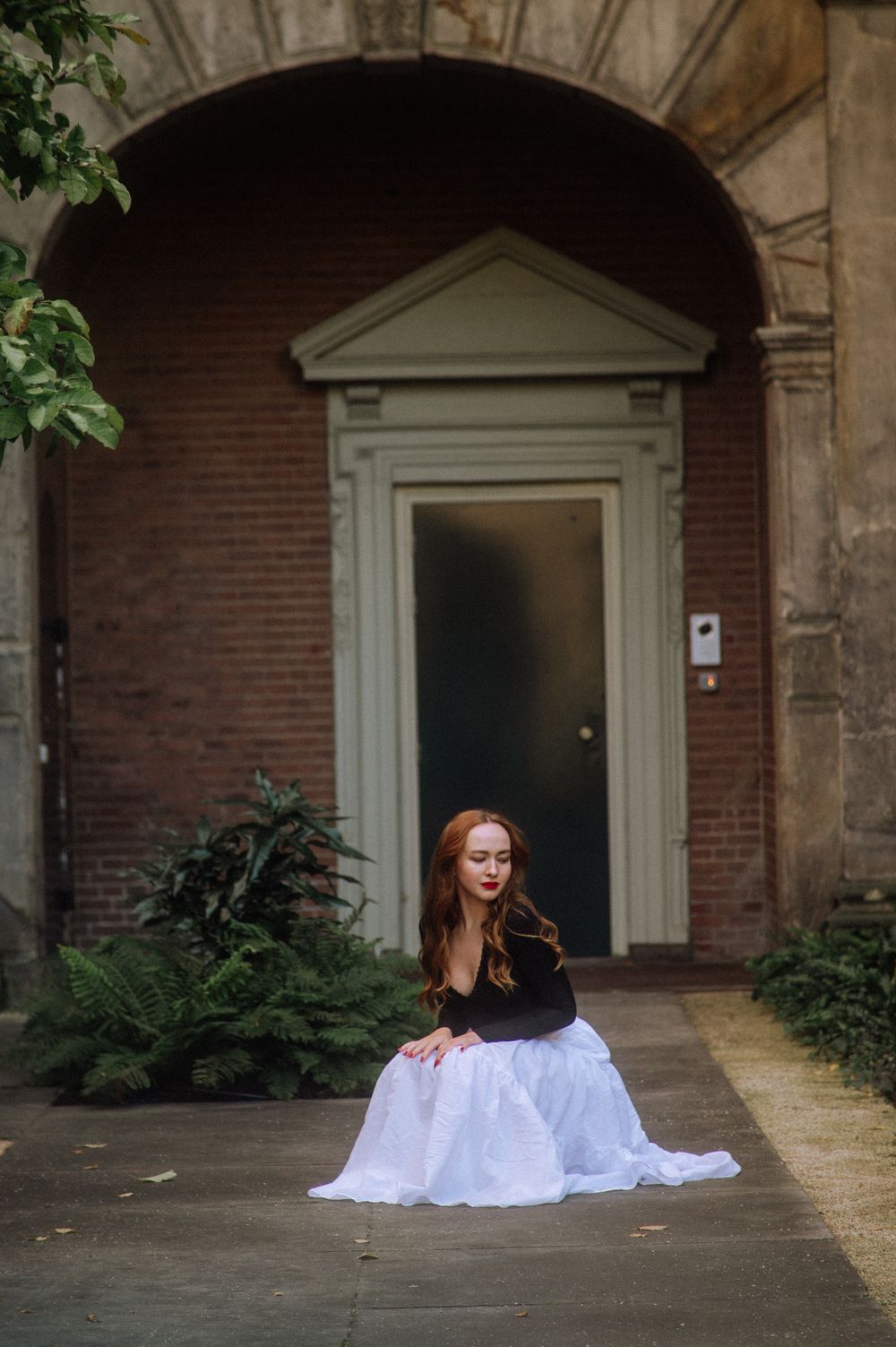 A person in a flowing white dress sits in front of arched doorways and brick architecture.