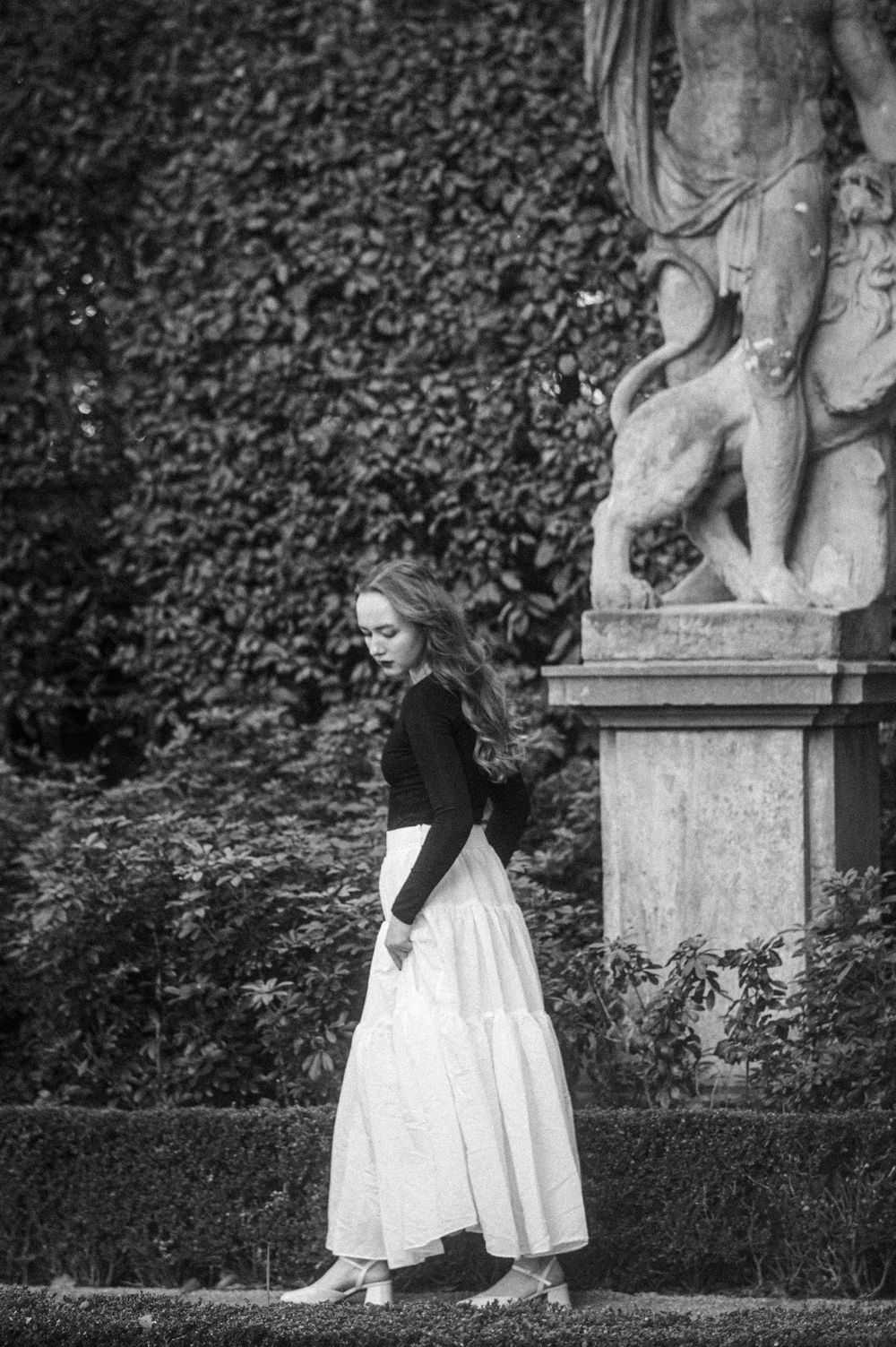 A figure in a black and white outfit poses near garden statues in a dramatic black and white photo.