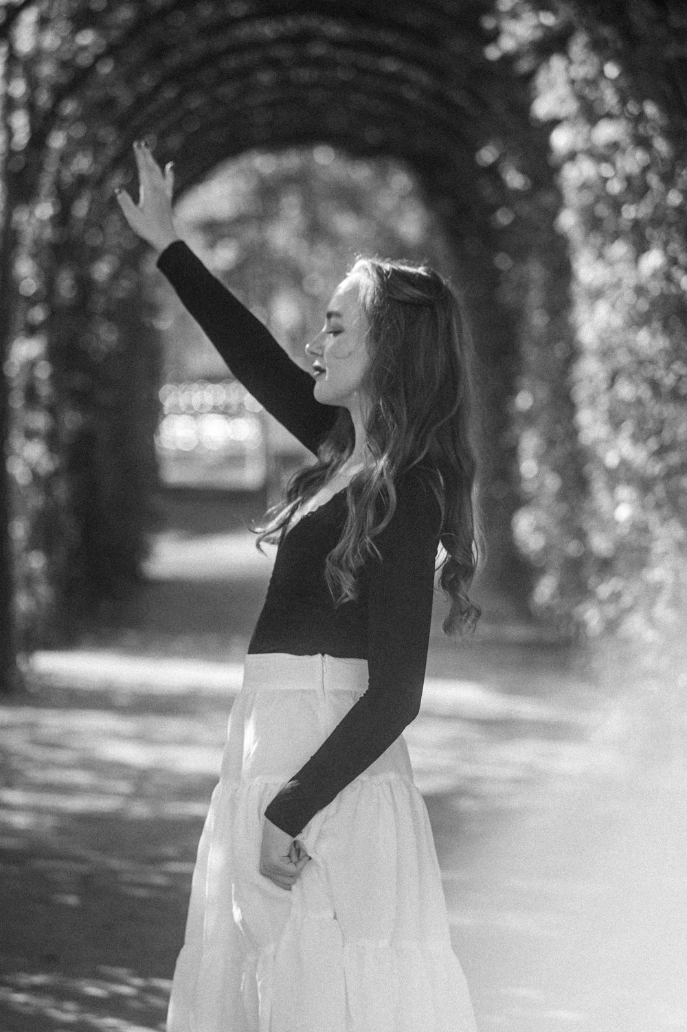 A person in a white dress and black top dances joyfully in a sunlit garden pathway.