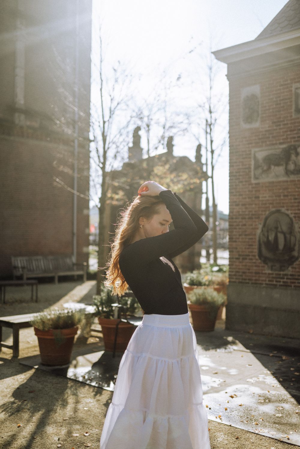 A figure in black and white clothing poses in sunlit courtyard with potted plants.