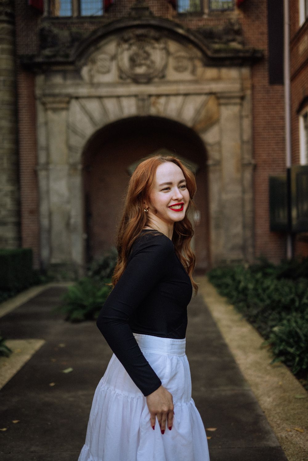 A smiling red-haired woman in the gardens of the Rijksmuseum with historic architecture in the background.