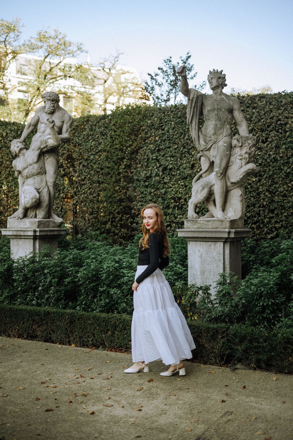 A figure in a white skirt and black top poses between classical statues in a formal garden setting.