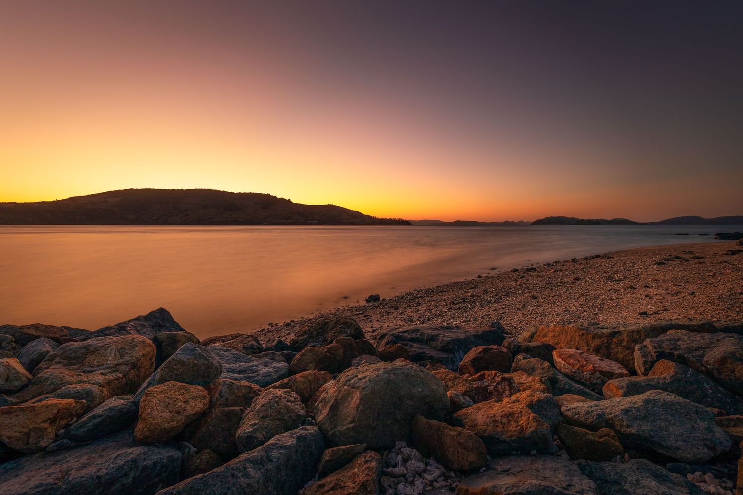 Rocky coastline silhouetted against golden sunset sky with mountain ridges in the distance.