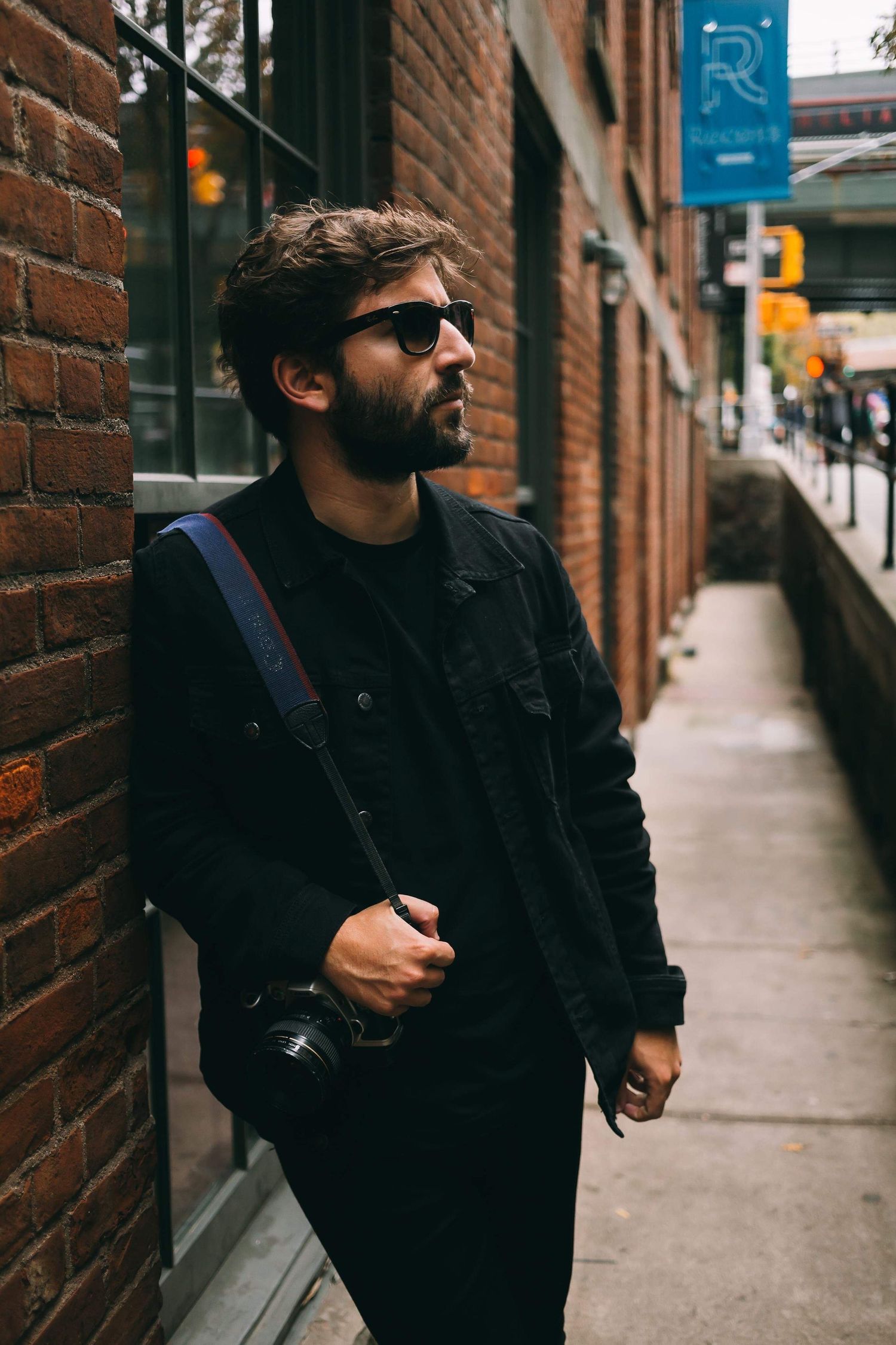 Street photographer in dark clothing carrying camera gear along brick building sidewalk.