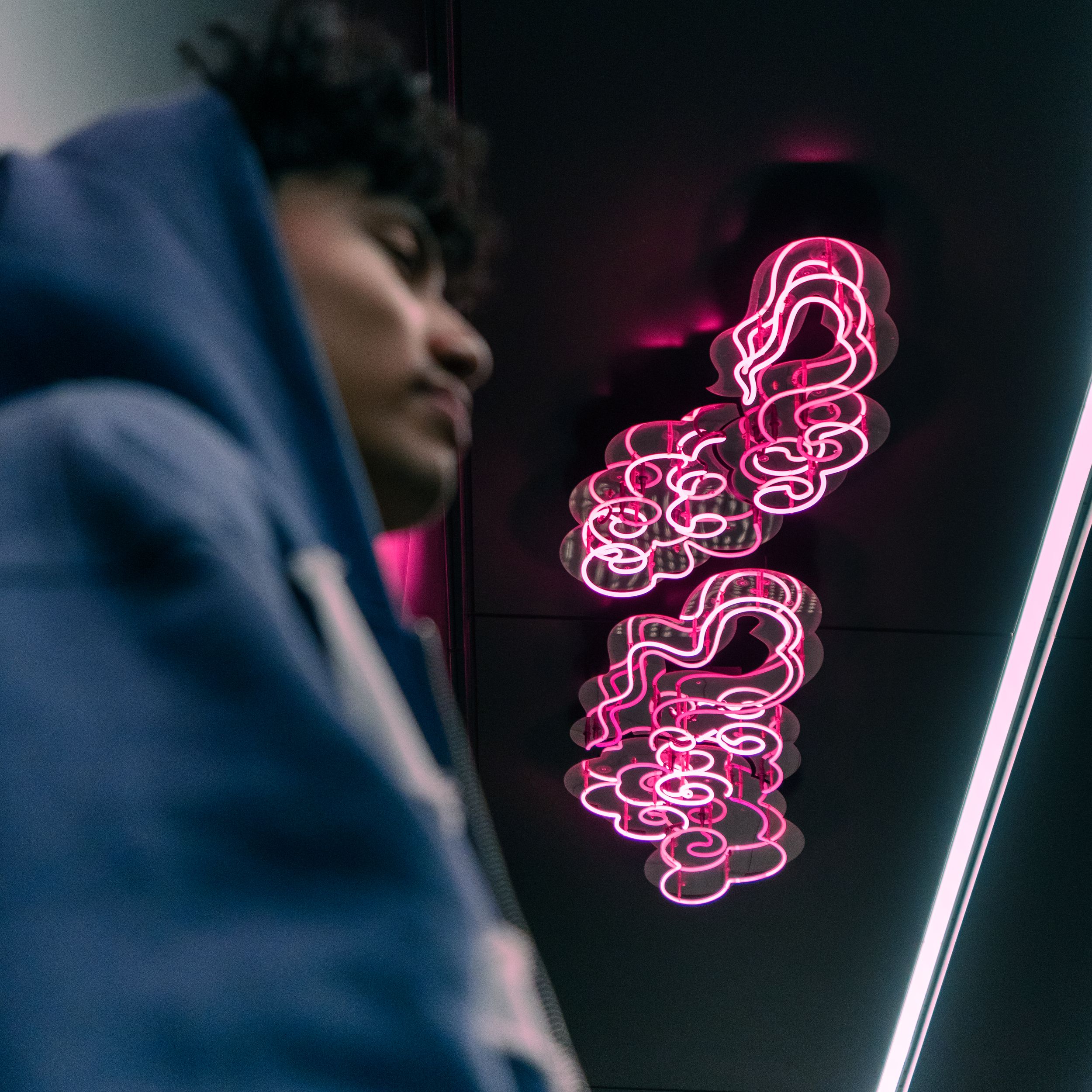 Man in blue hoodie standing beside pink neon light sign in dark moody indoor setting.