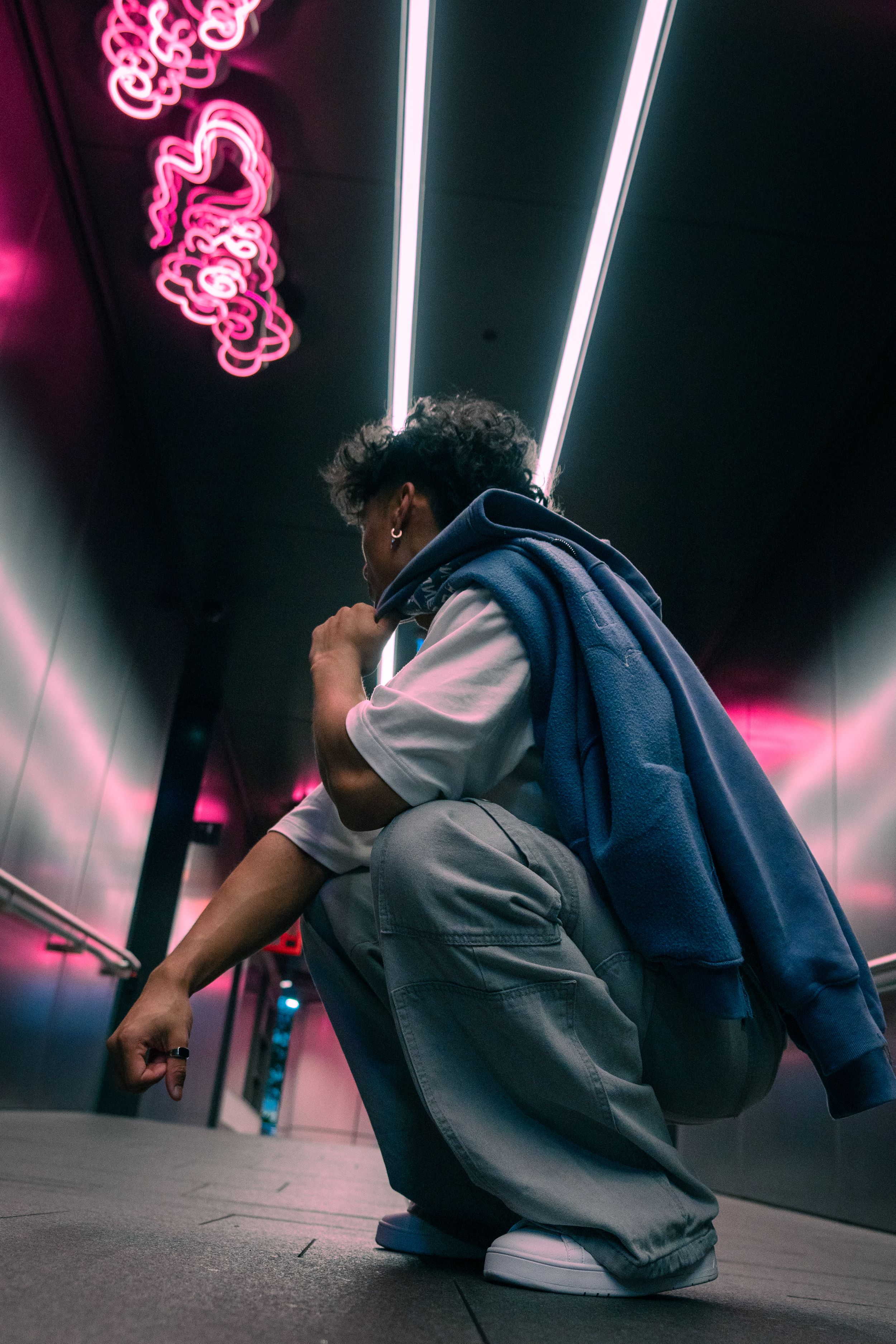 Young man crouching in white shirt and grey pants under pink neon lights in dark corridor.
