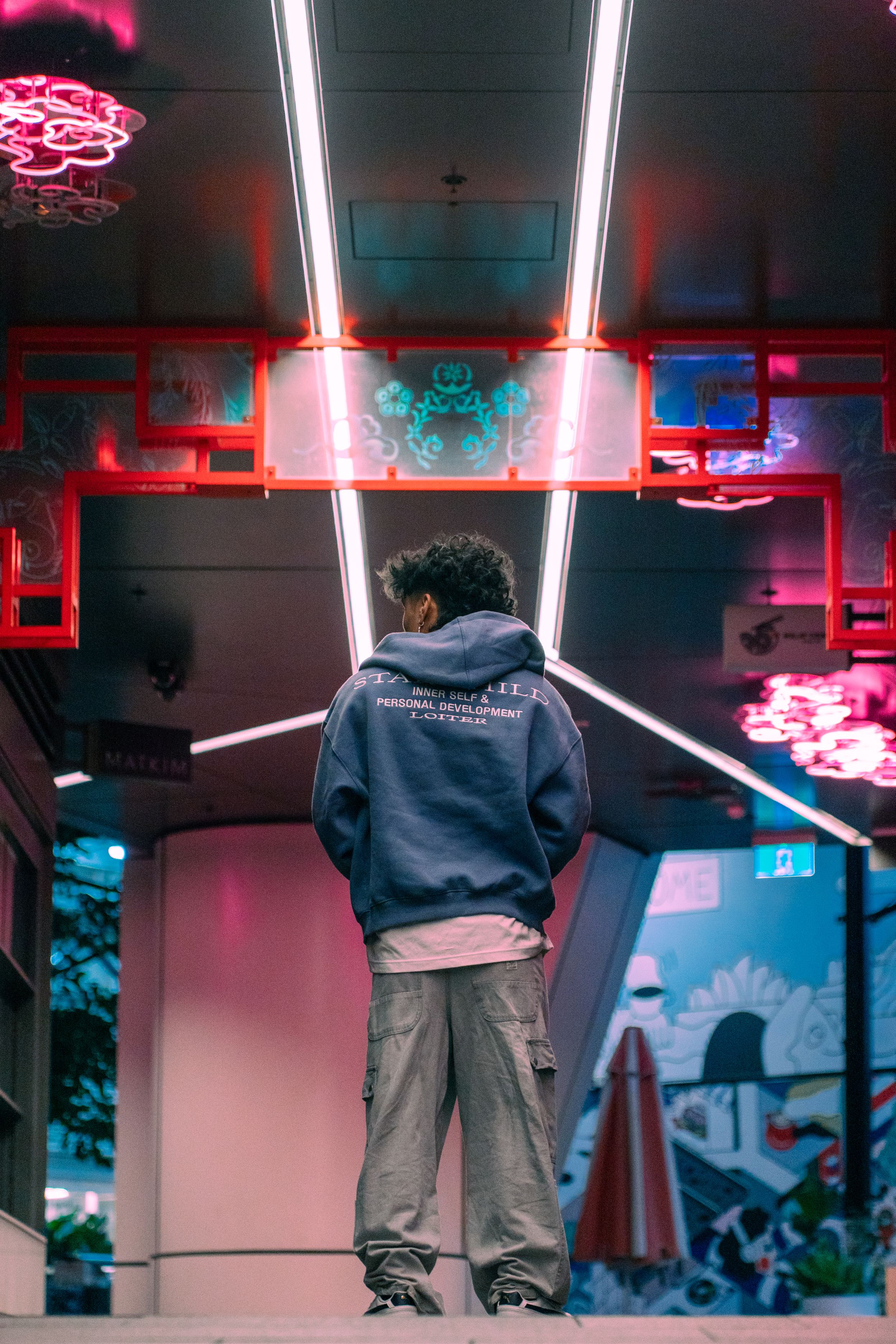Person in hoodie and cargo pants standing under neon lights at night.