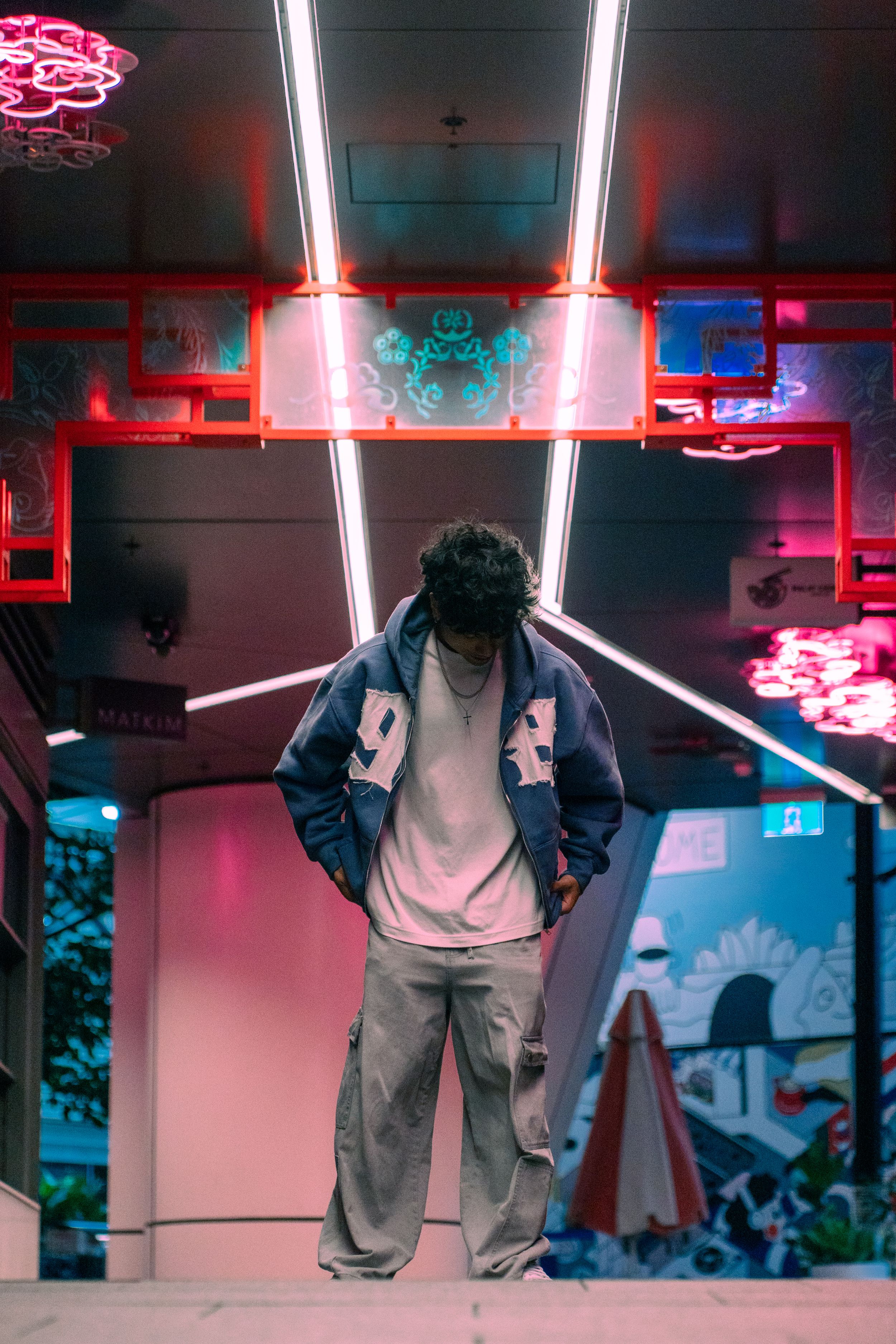 Man in varsity jacket posing under neon lights at colorful night spot.