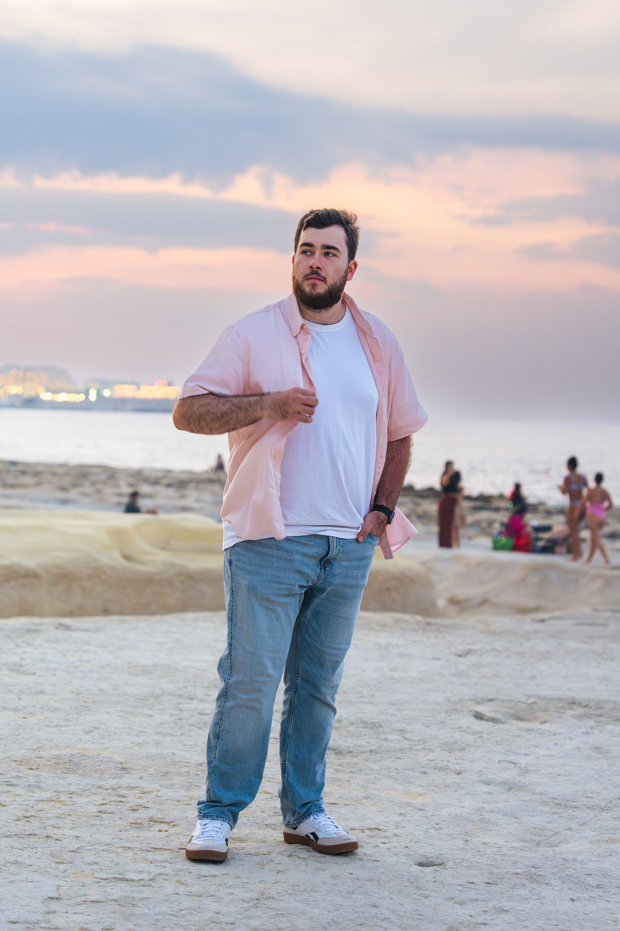 Young bearded man in pink shirt and jeans posing on rocky beach during colorful sunset with people in background.