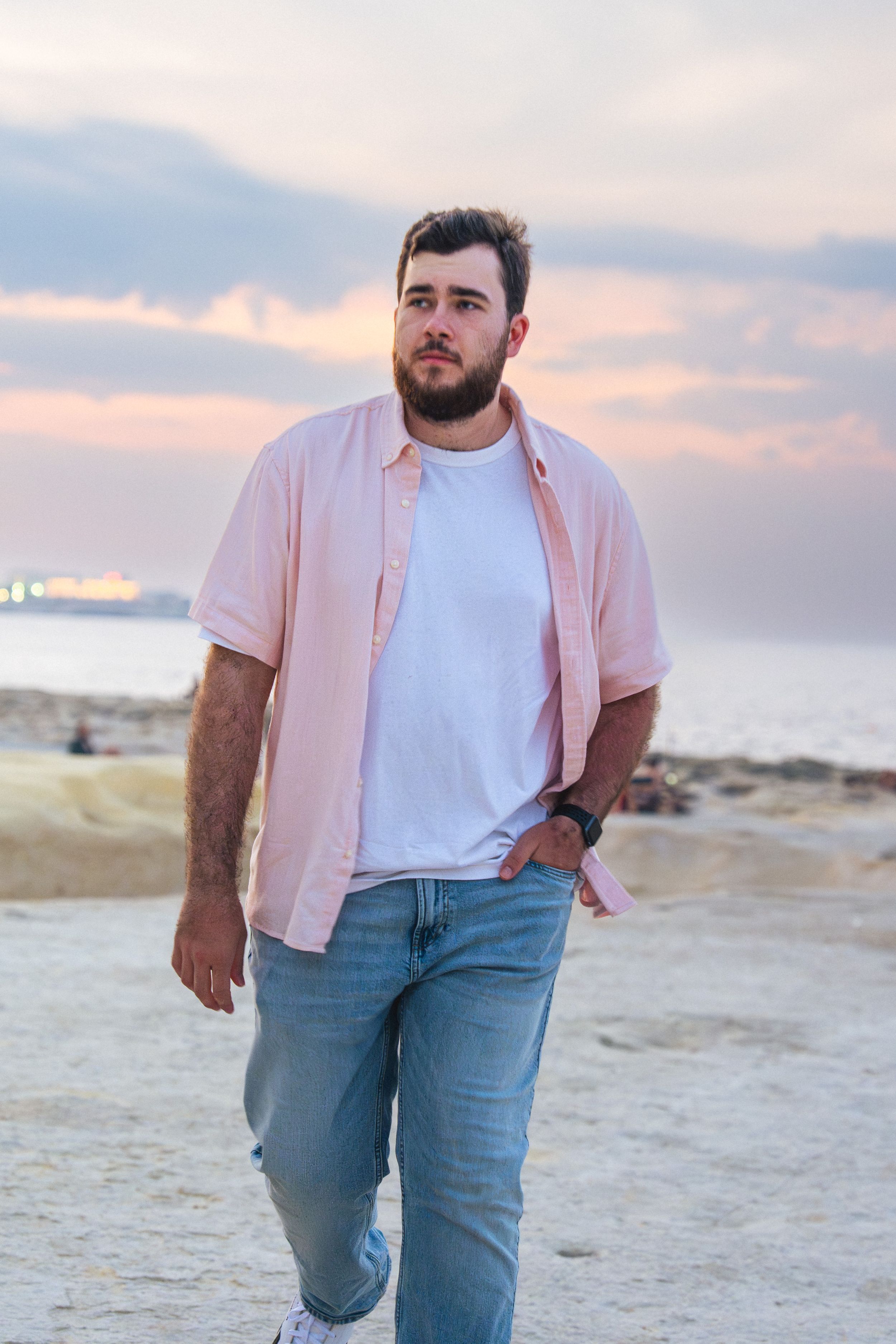 Bearded man in pink shirt and jeans walking on rocky beach at sunset with ocean in background.