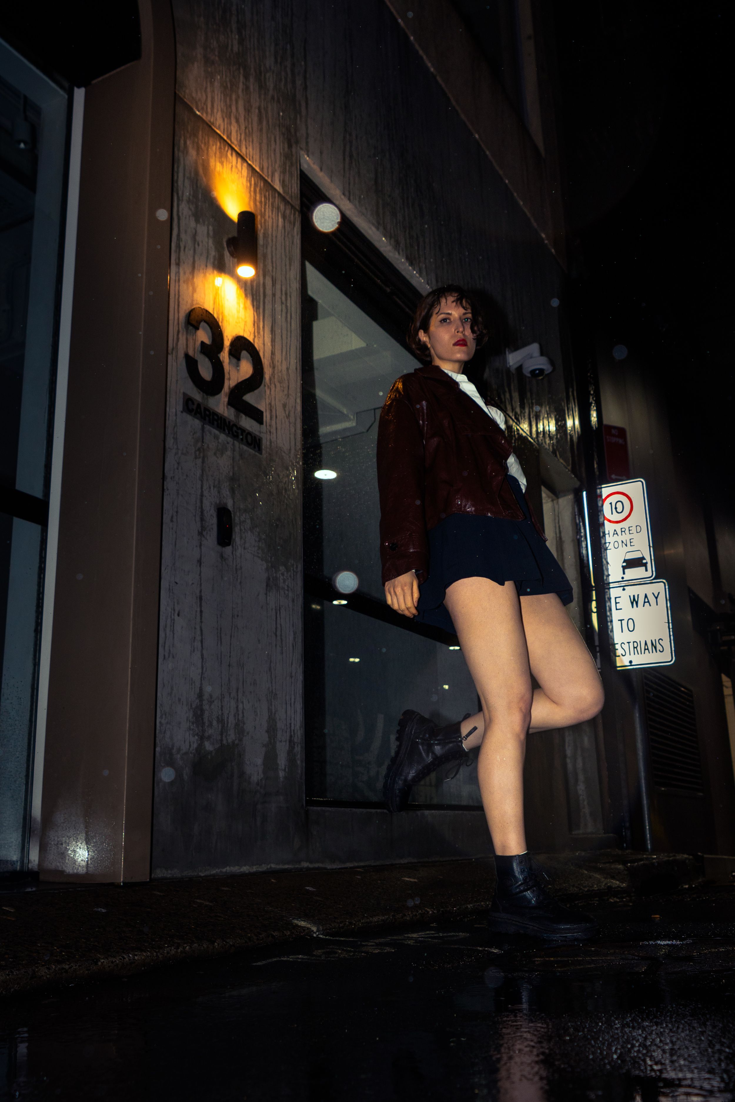 Woman in red jacket and blue mini skirt posing at night outside building number 32 on wet city street.