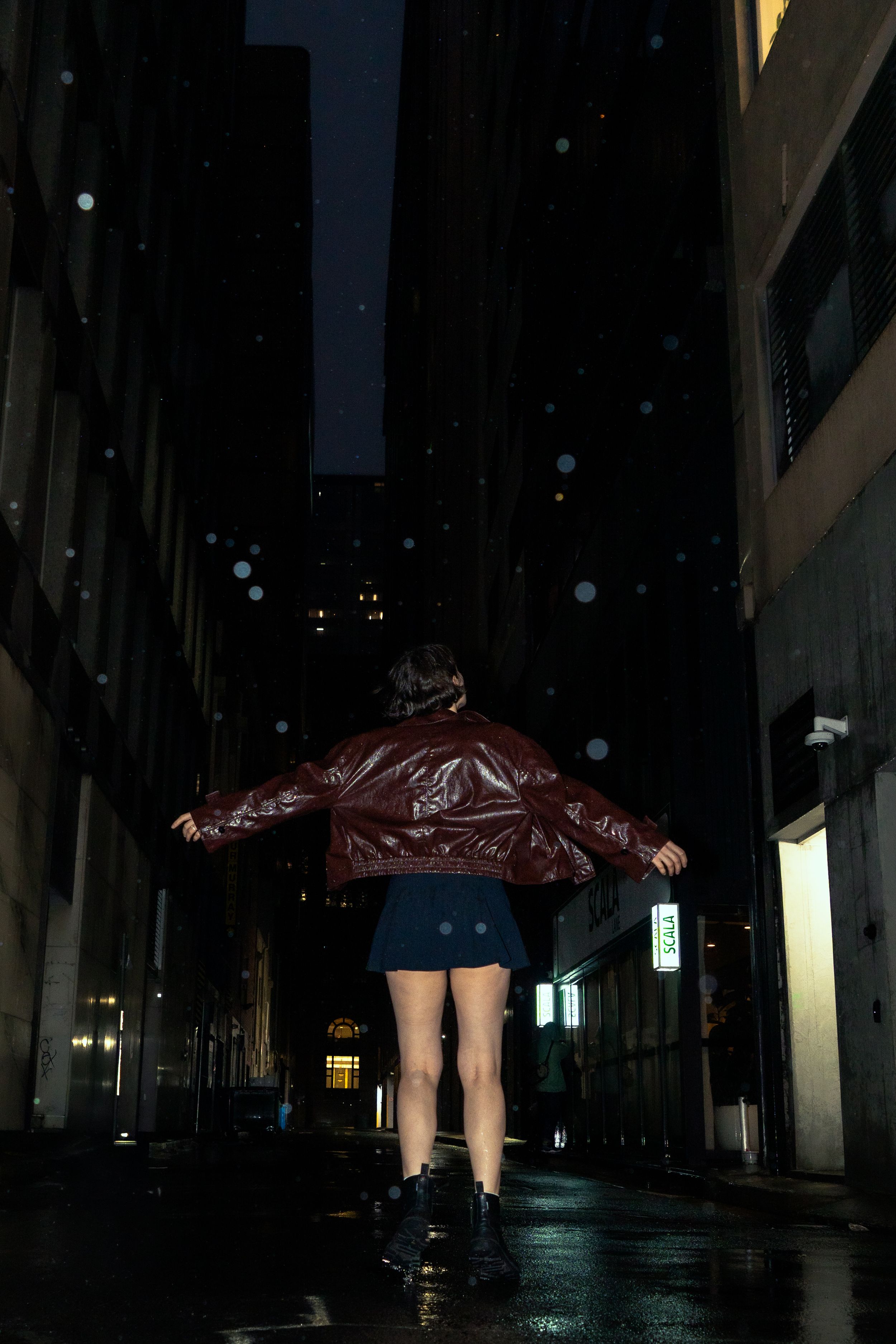 Woman in red leather jacket and blue mini skirt standing in rainy dark city alley at night.