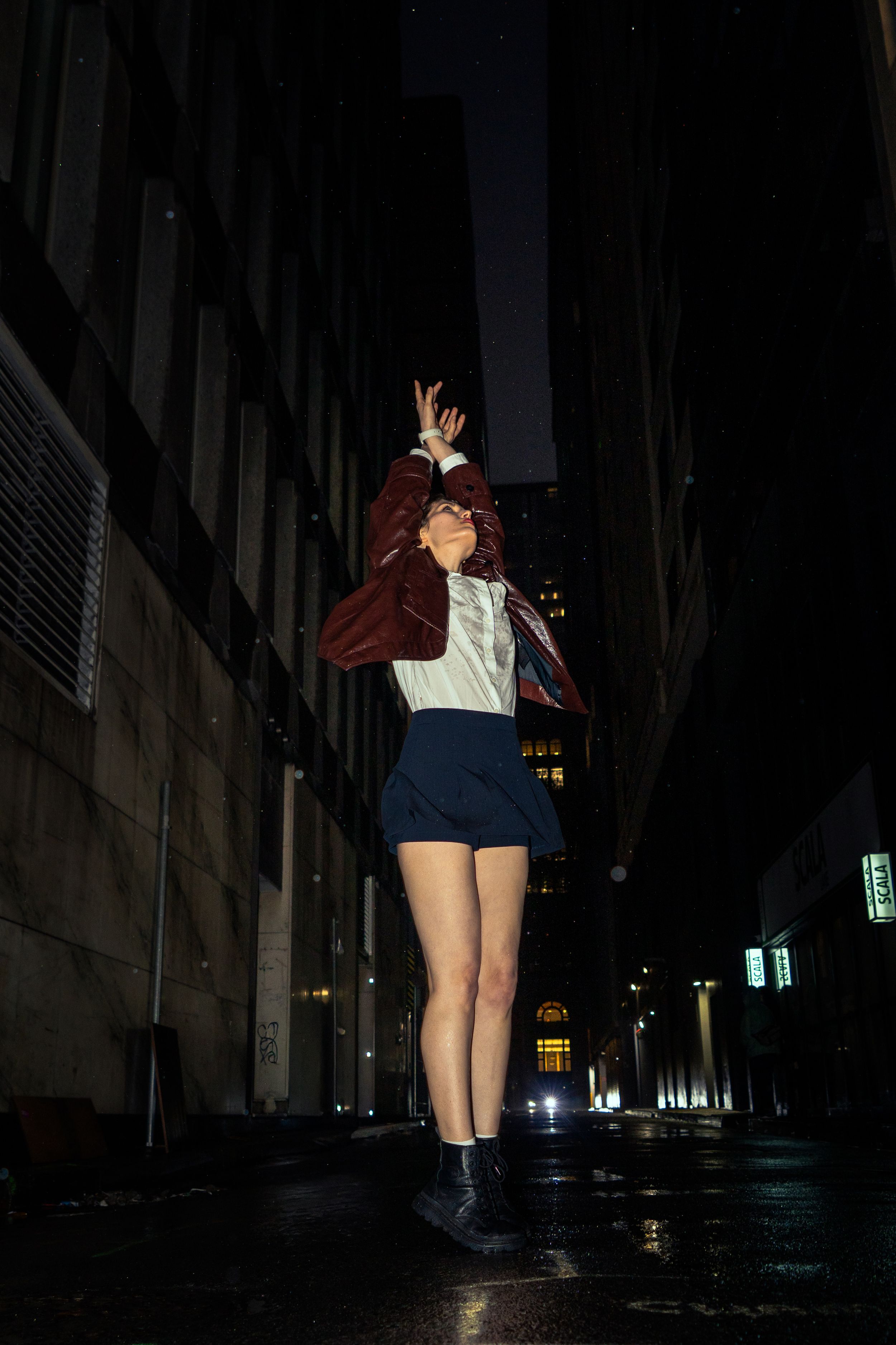 Woman in red jacket and blue skirt jumping with arms raised in dark wet city alley at night.