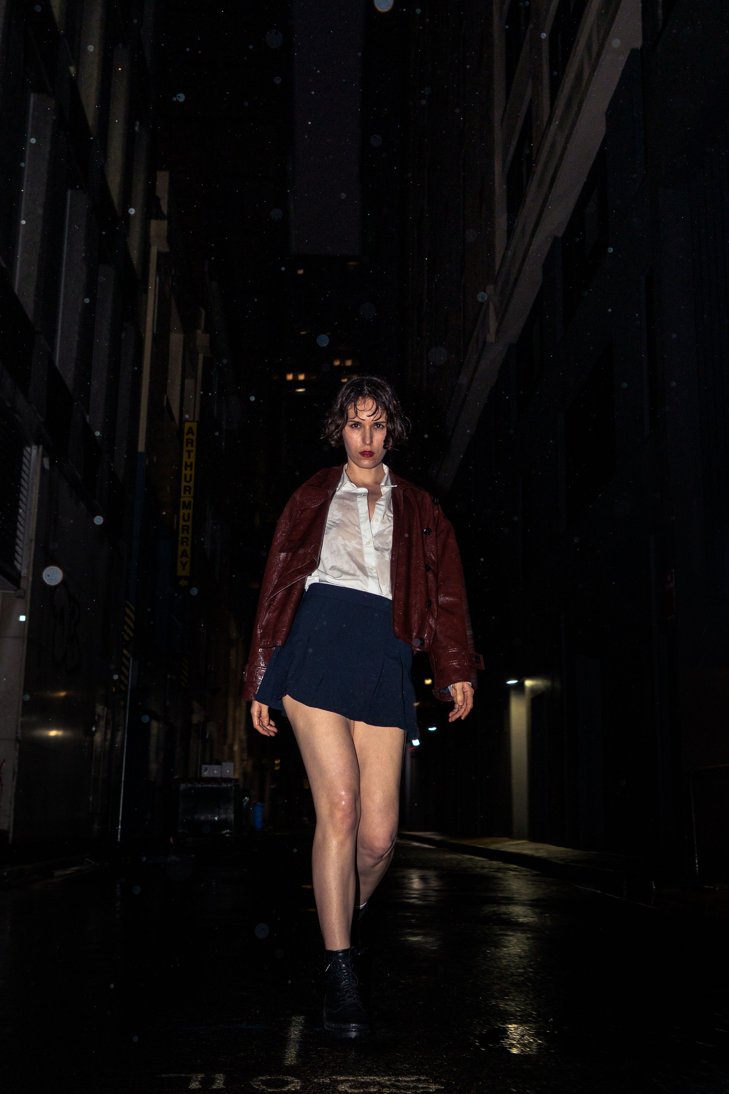 Woman in red jacket and blue mini skirt walking confidently through dark rainy city alley at night.