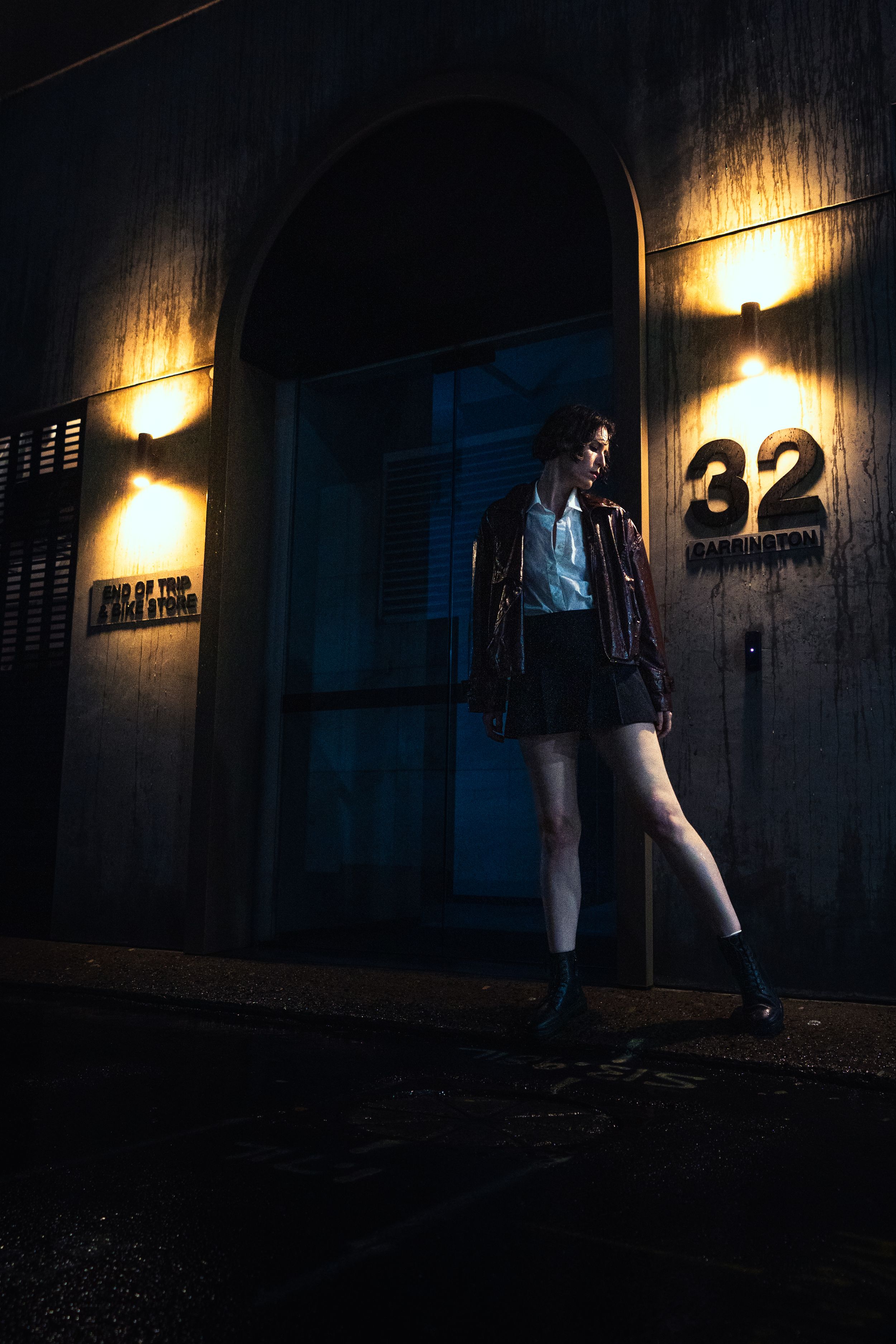 Woman in red leather jacket posing outside illuminated building number 32 on dark rainy night.