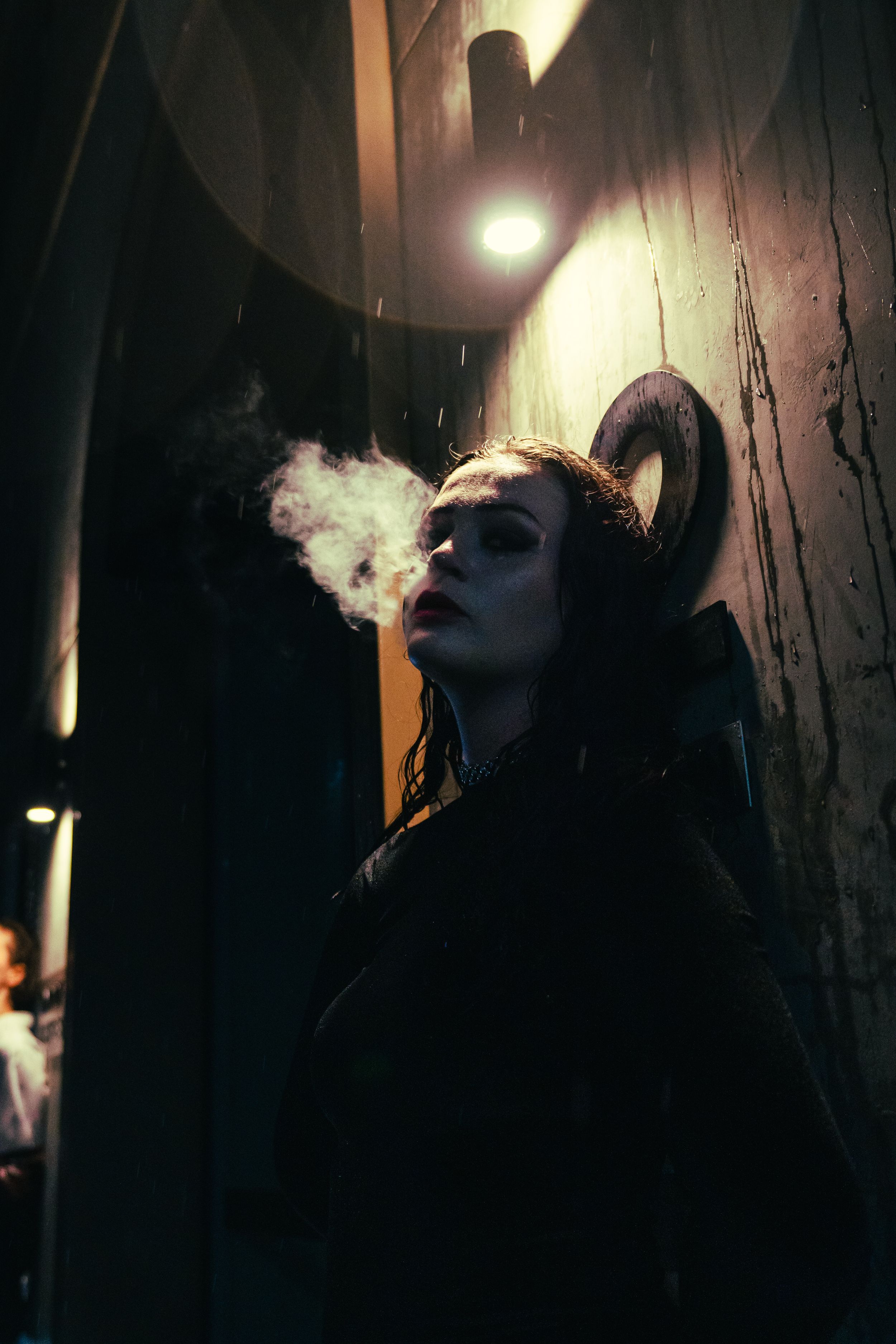 Woman wearing dark coat and ram horns exhaling smoke in dramatic moody dark alley at night.