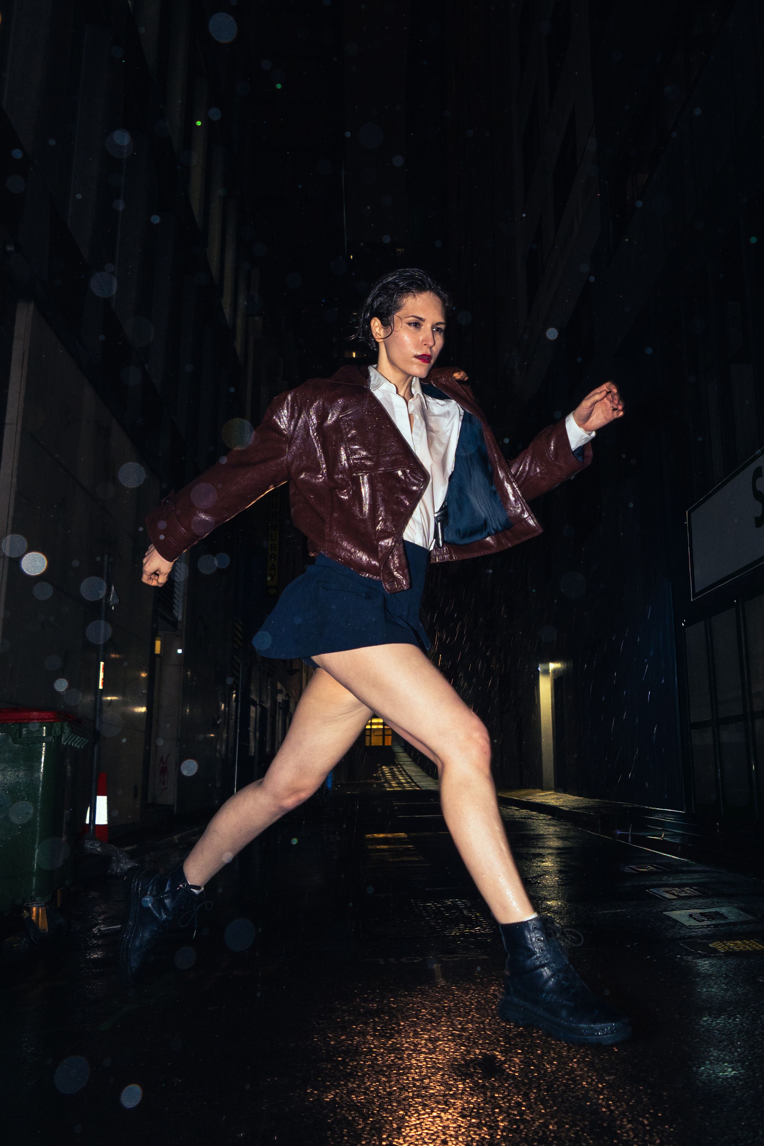 Woman in red jacket and blue mini skirt running dynamically through dark rainy city street at night.