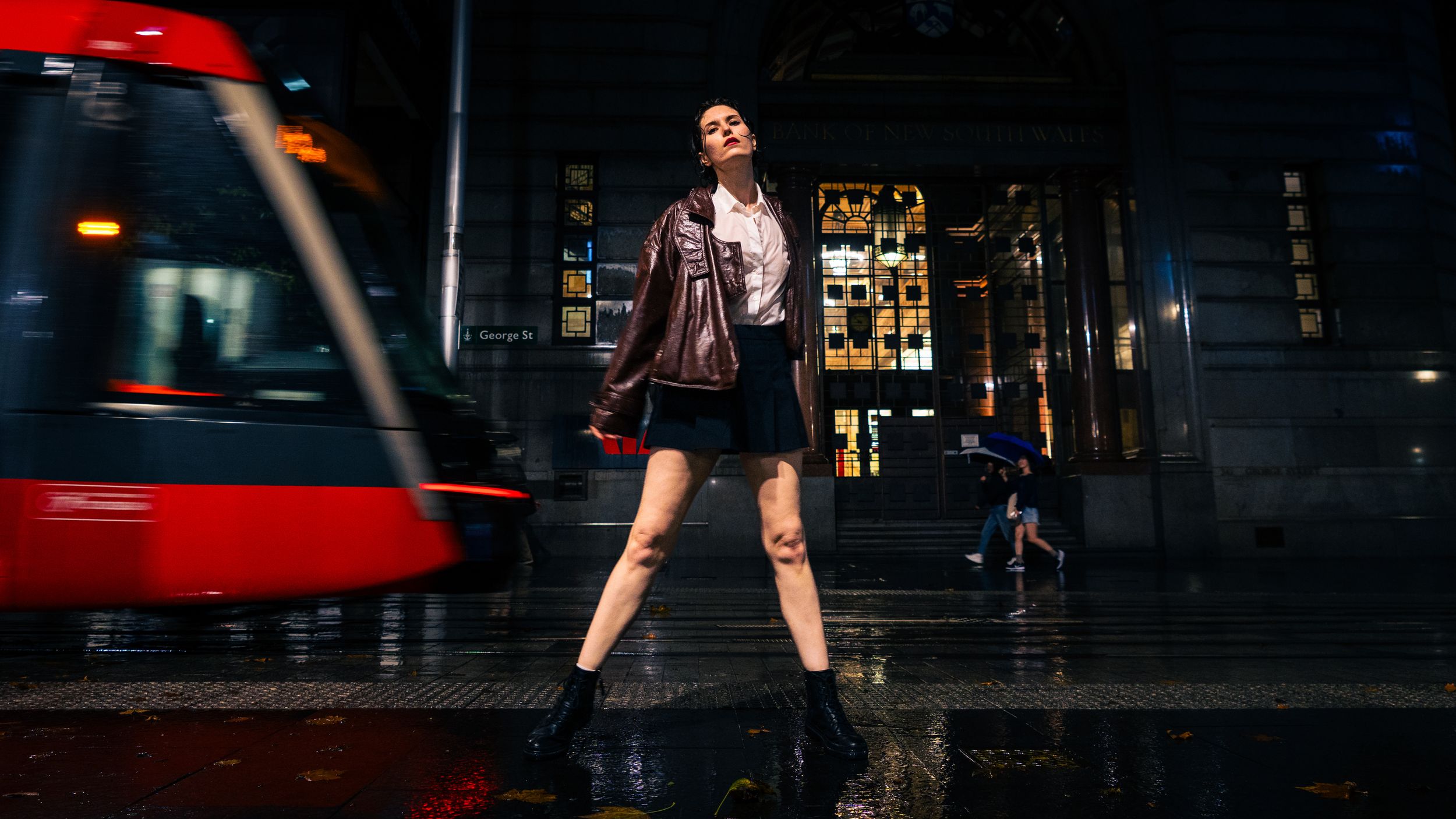 Young woman posing confidently on a rainy city street at night with a tram passing in the background.