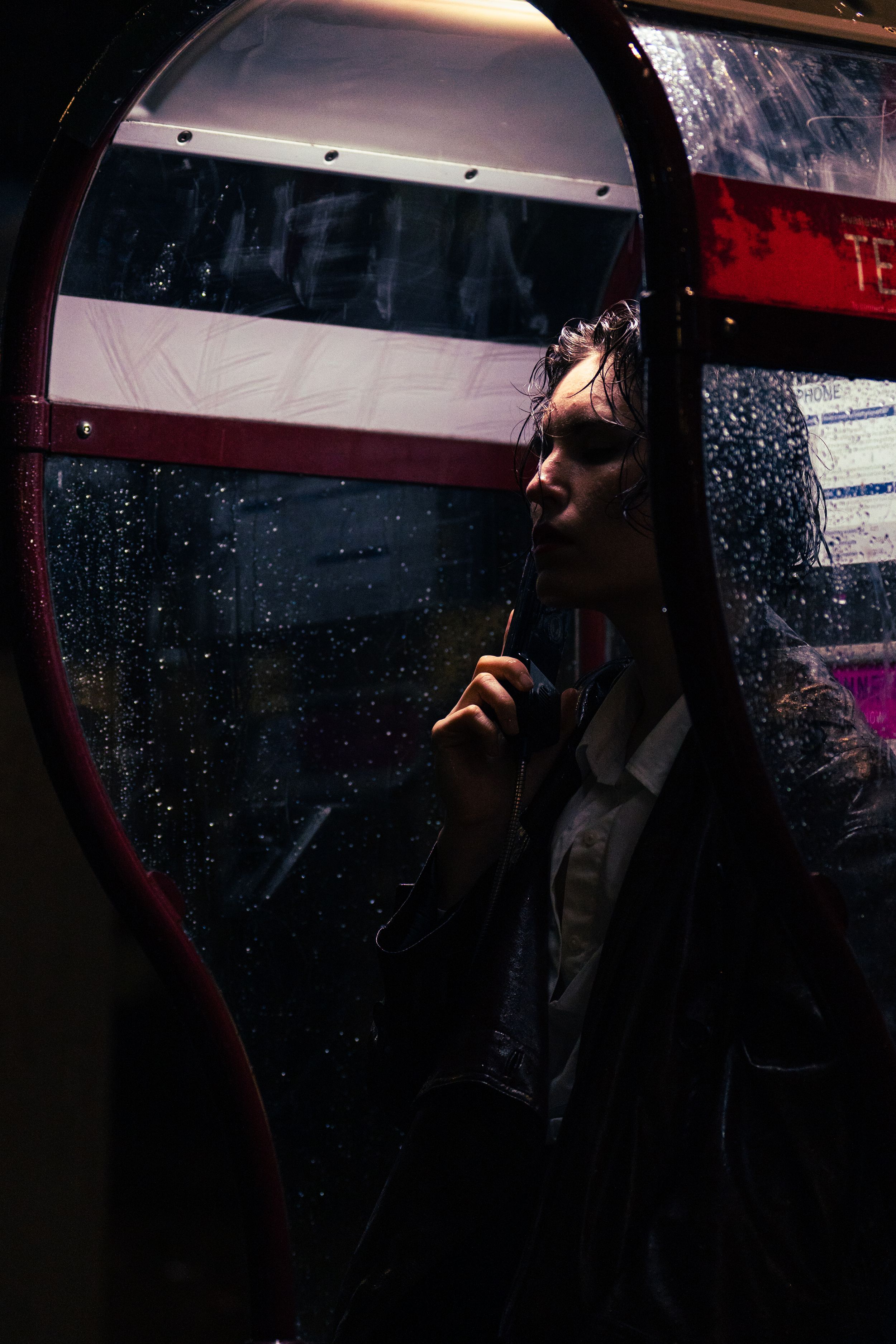 Person inside a rain-covered phone booth at night, holding a cigarette in moody dramatic lighting.