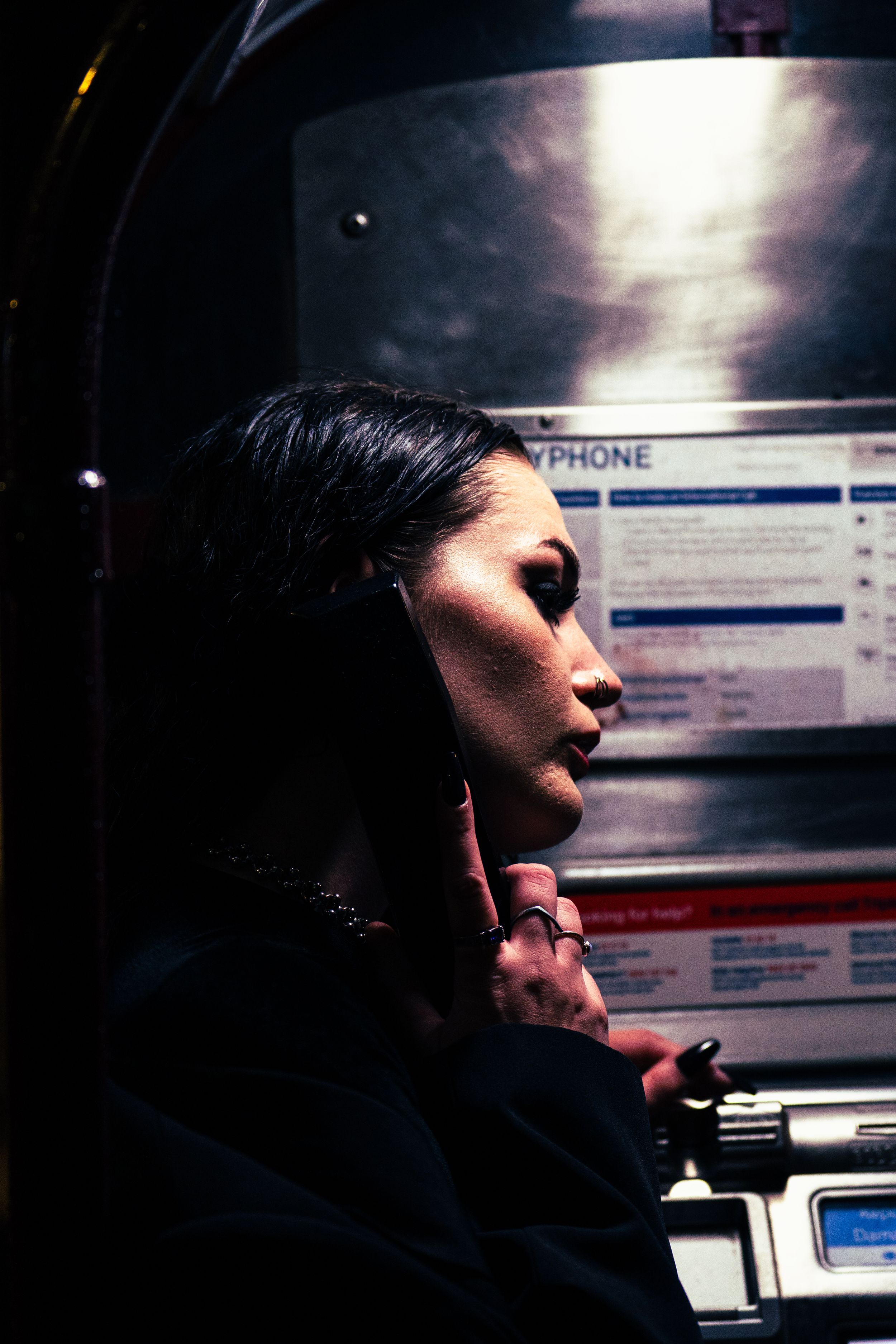 Woman using a payphone at night, dramatic side lighting highlighting her profile in urban setting.