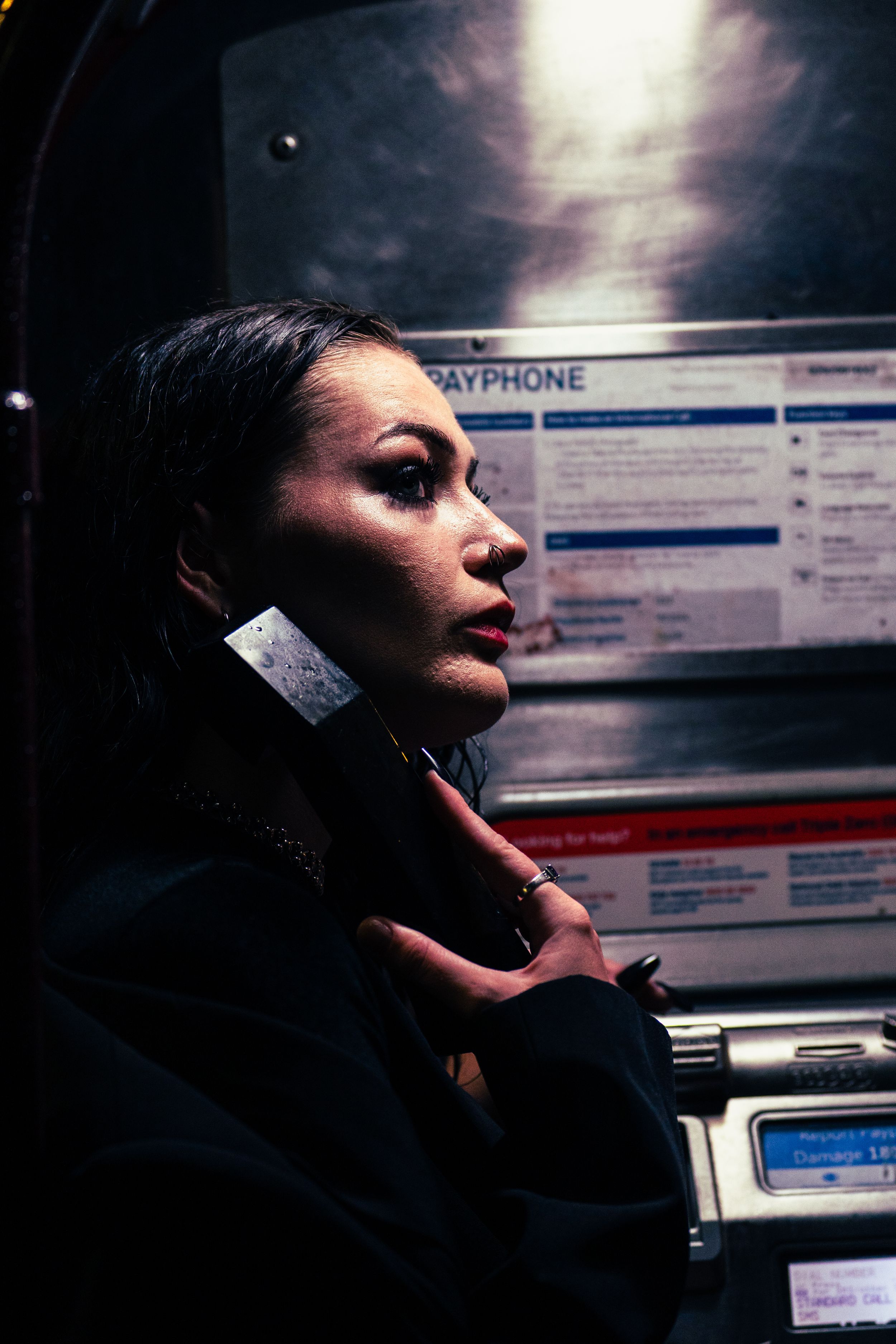 Woman on a payphone at night, dramatic lighting illuminating her face against dark background.