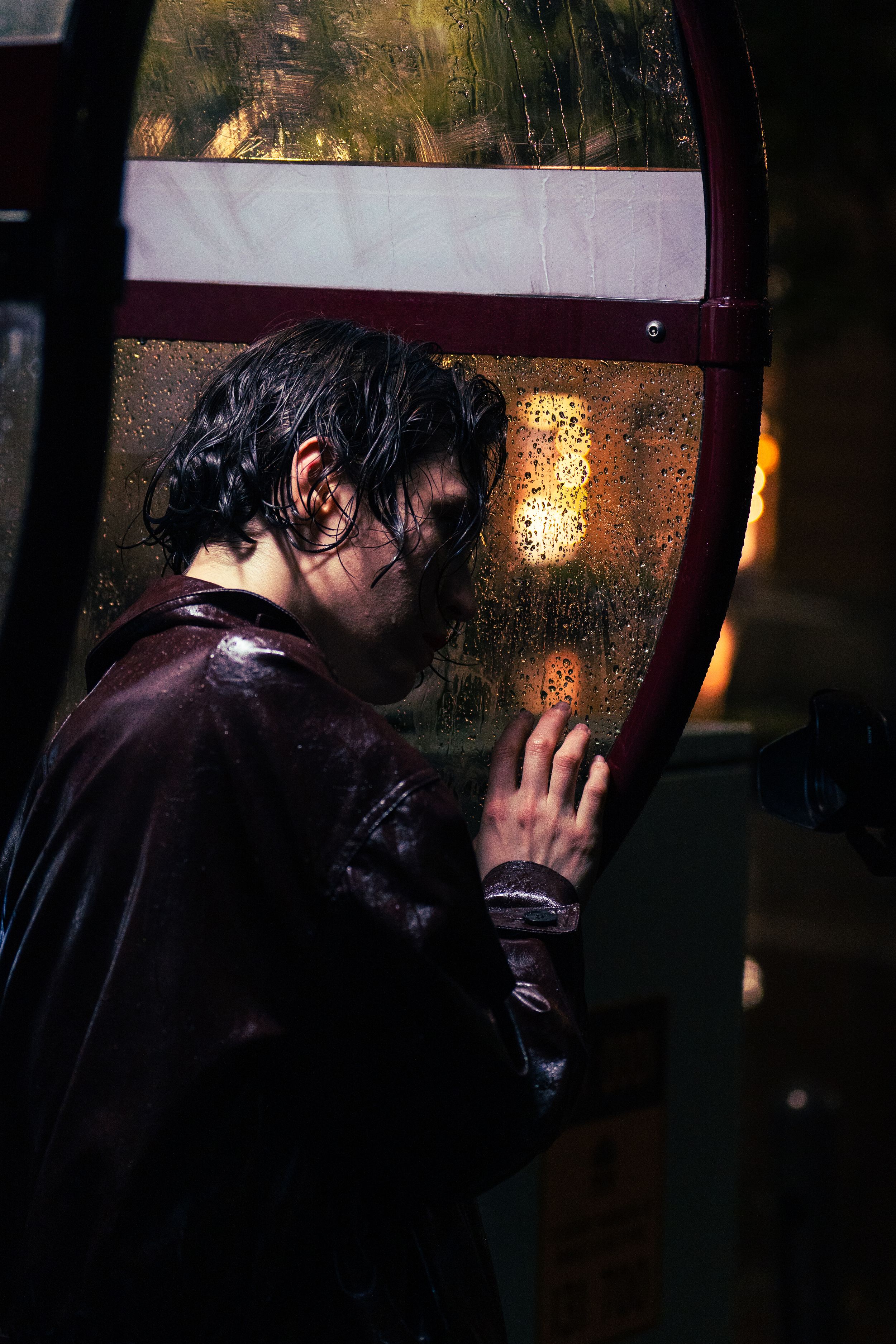 Person leaning against rain-soaked phone booth window at night, hand pressed against wet glass.