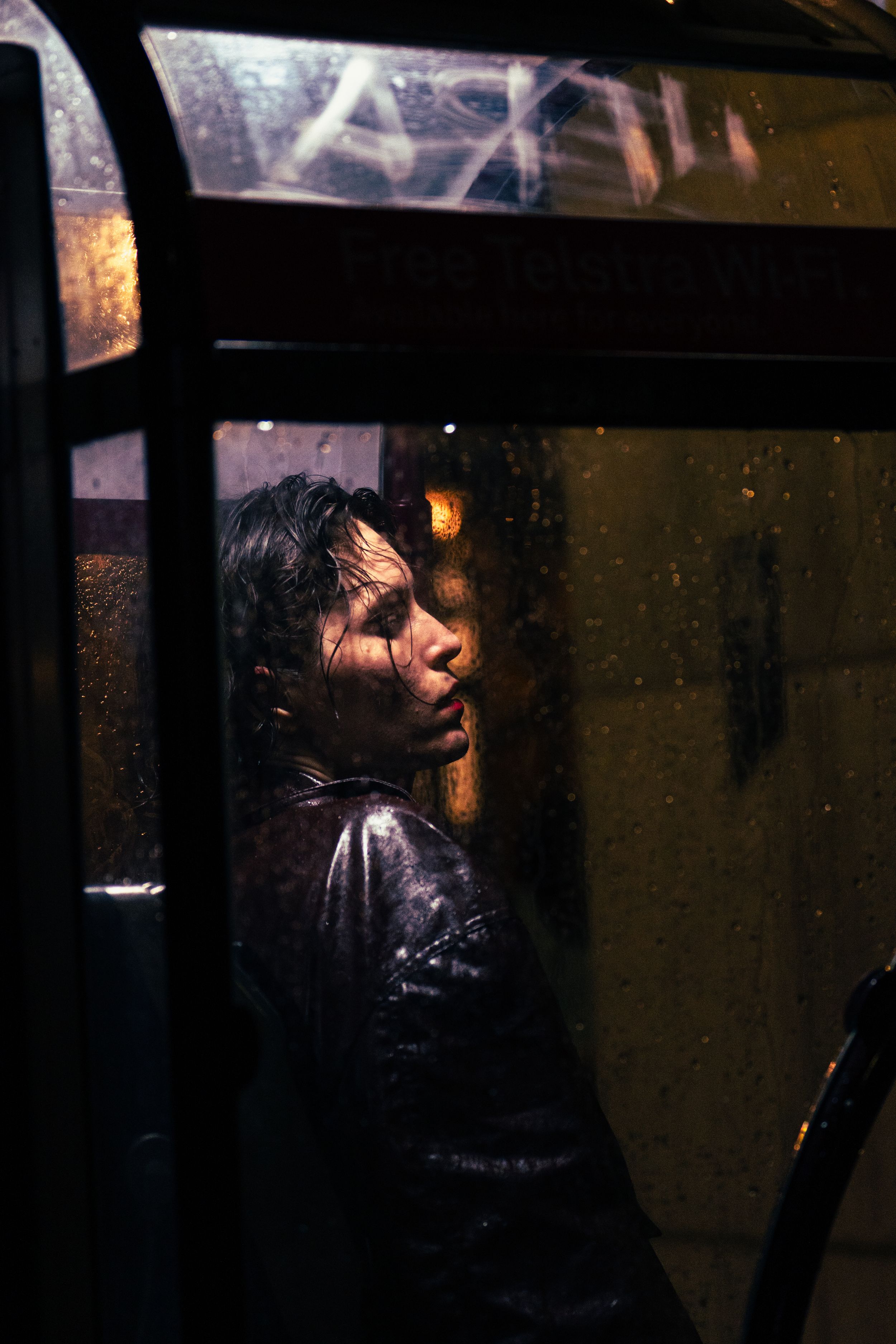 Person with wet hair seen through rain-covered bus window at night, moody urban photography.