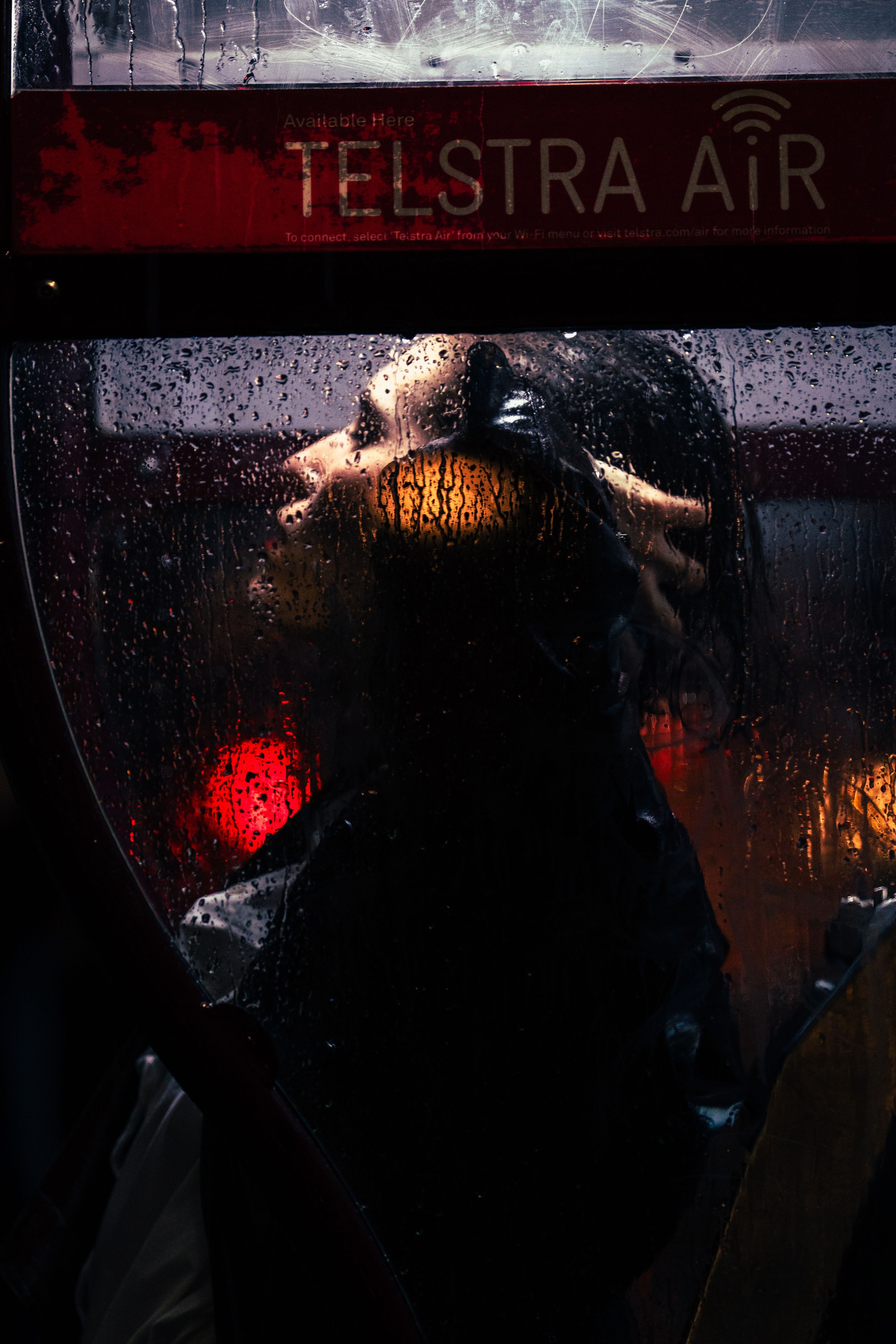 Silhouette of person inside Telstra Air phone booth, viewed through rain-covered glass at night.