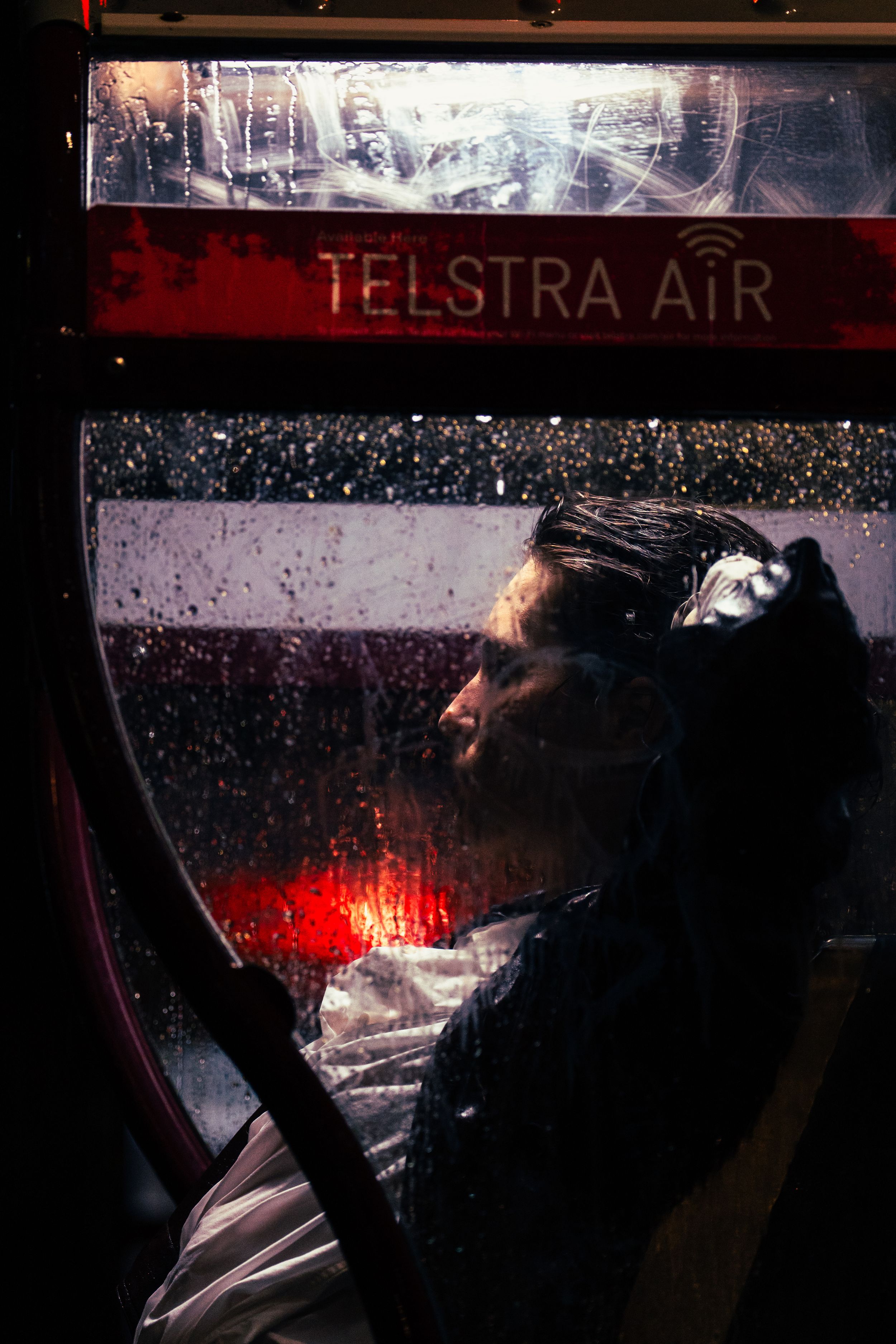 Person inside Telstra Air phone booth at night, face illuminated through rain-speckled glass.