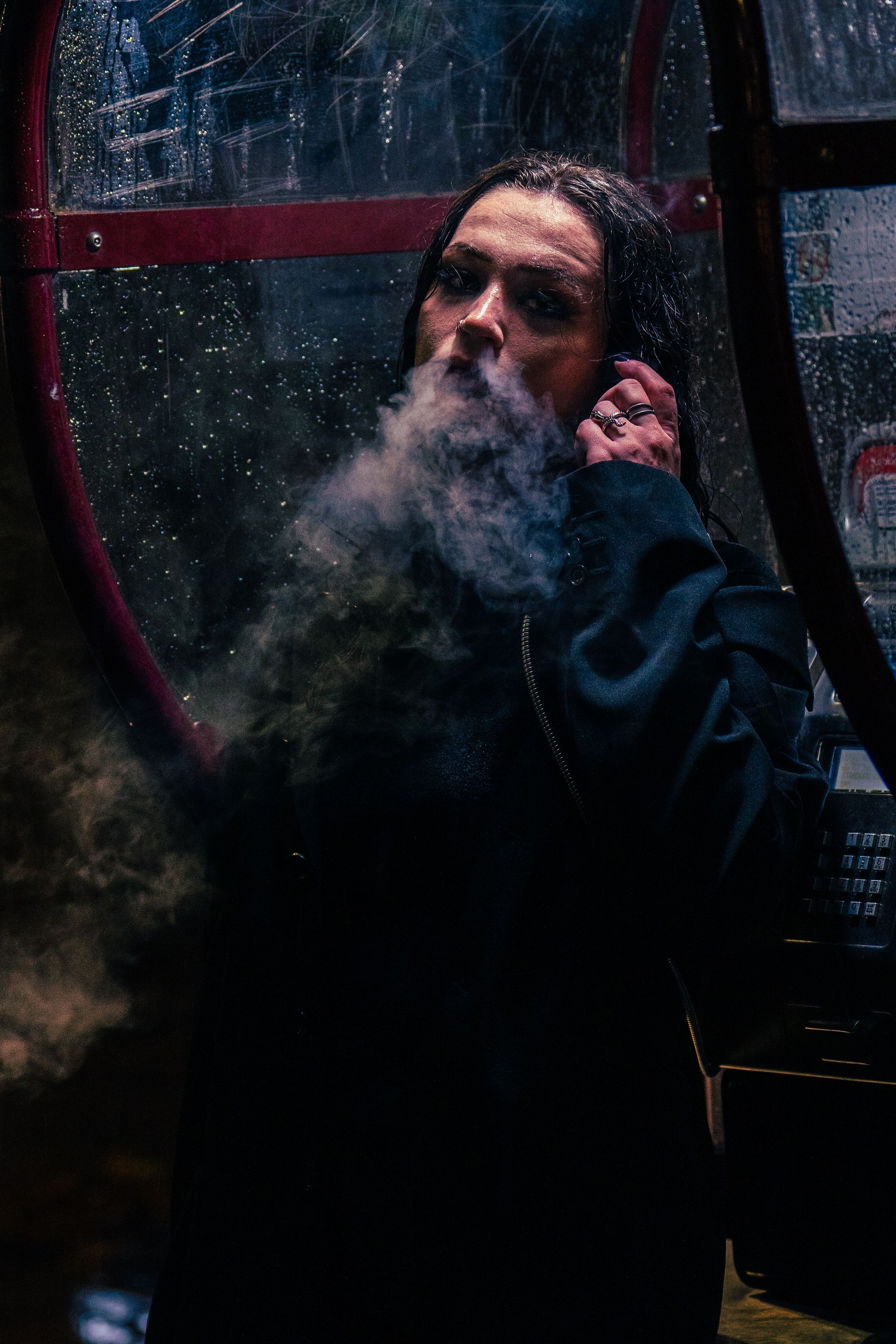 Person exhaling smoke outside a rain-covered phone booth at night in dramatic urban lighting.
