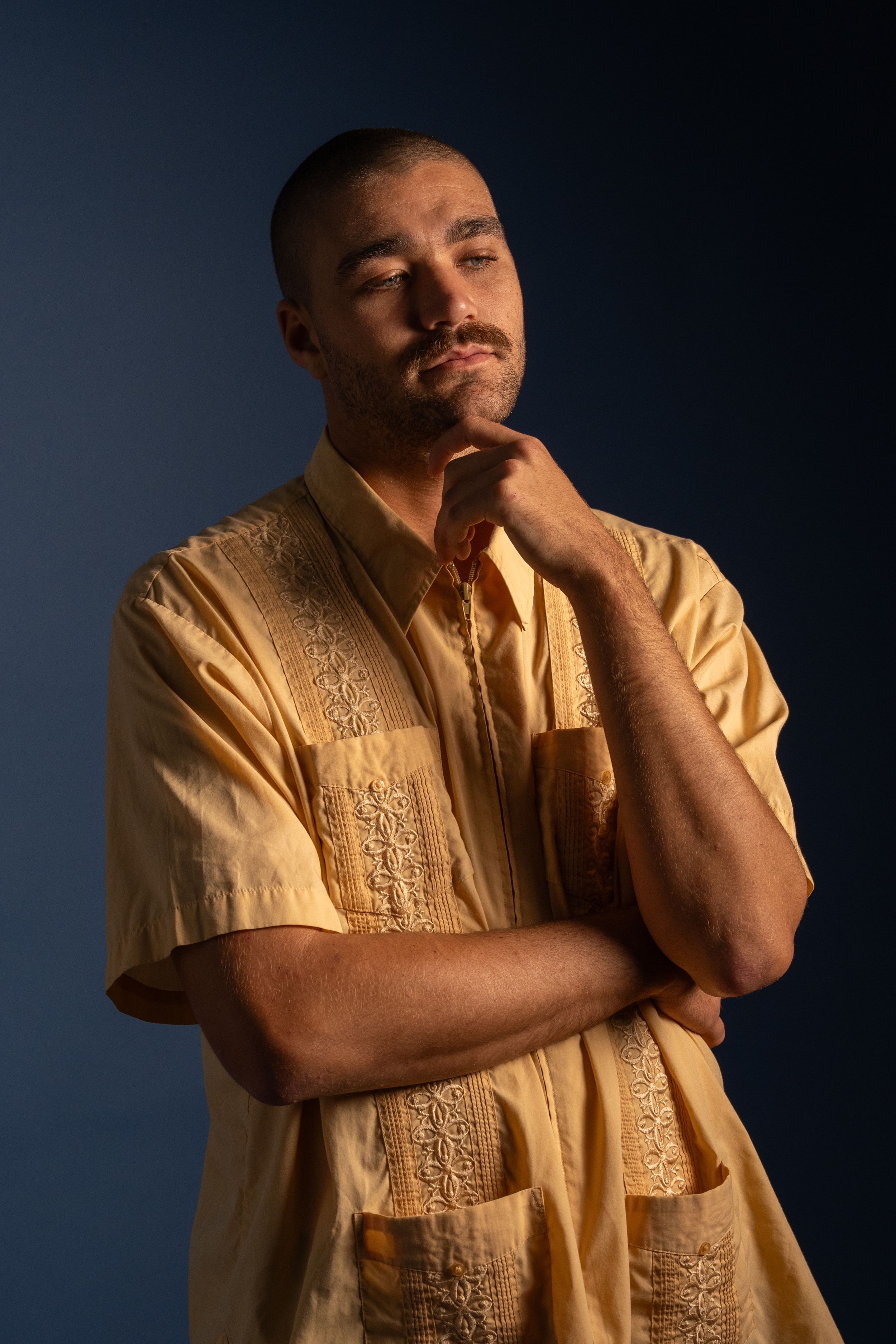 Man in yellow embroidered guayabera shirt posing thoughtfully against a dark blue background.