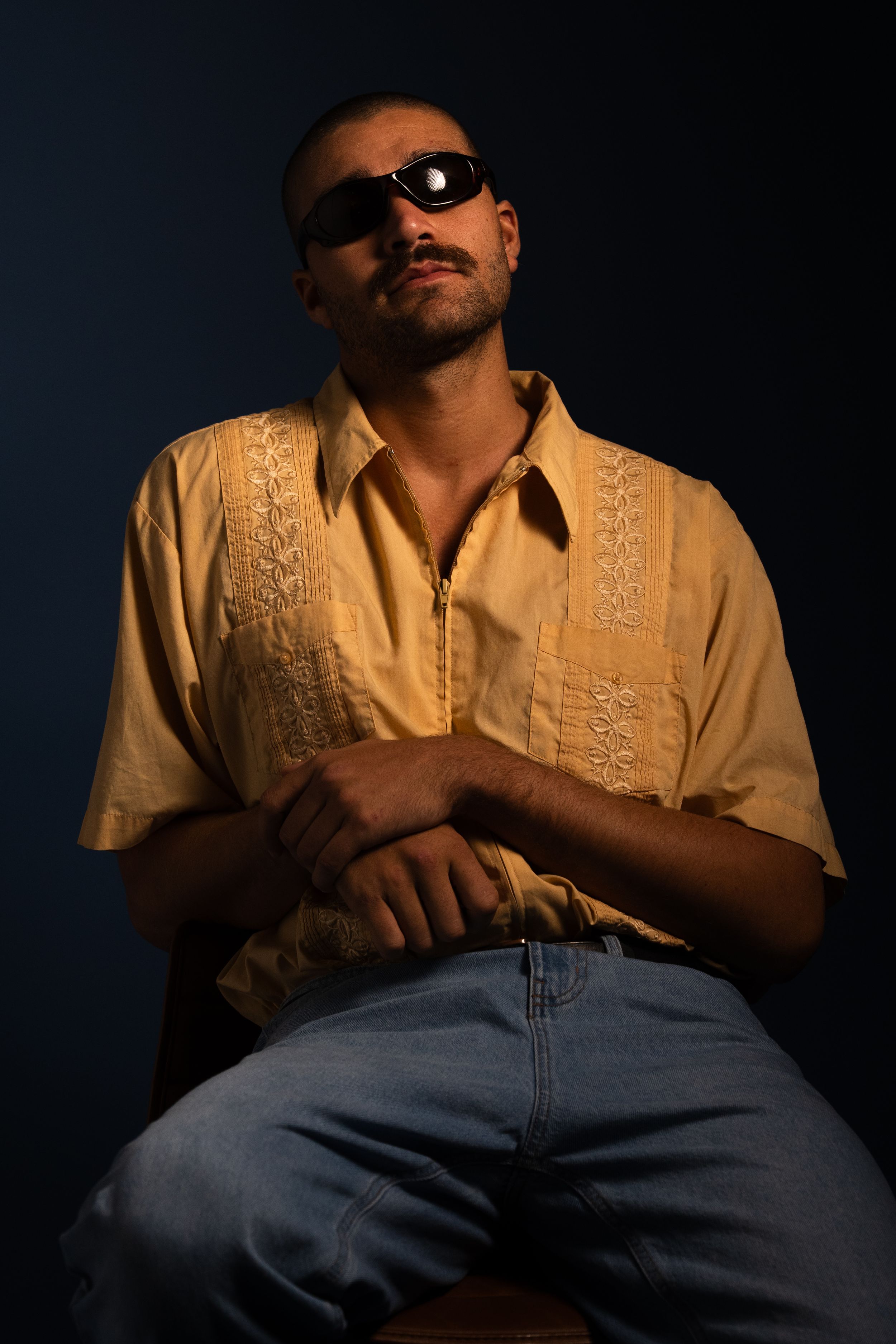 Man in yellow shirt and sunglasses sitting confidently against a dark dramatic background.