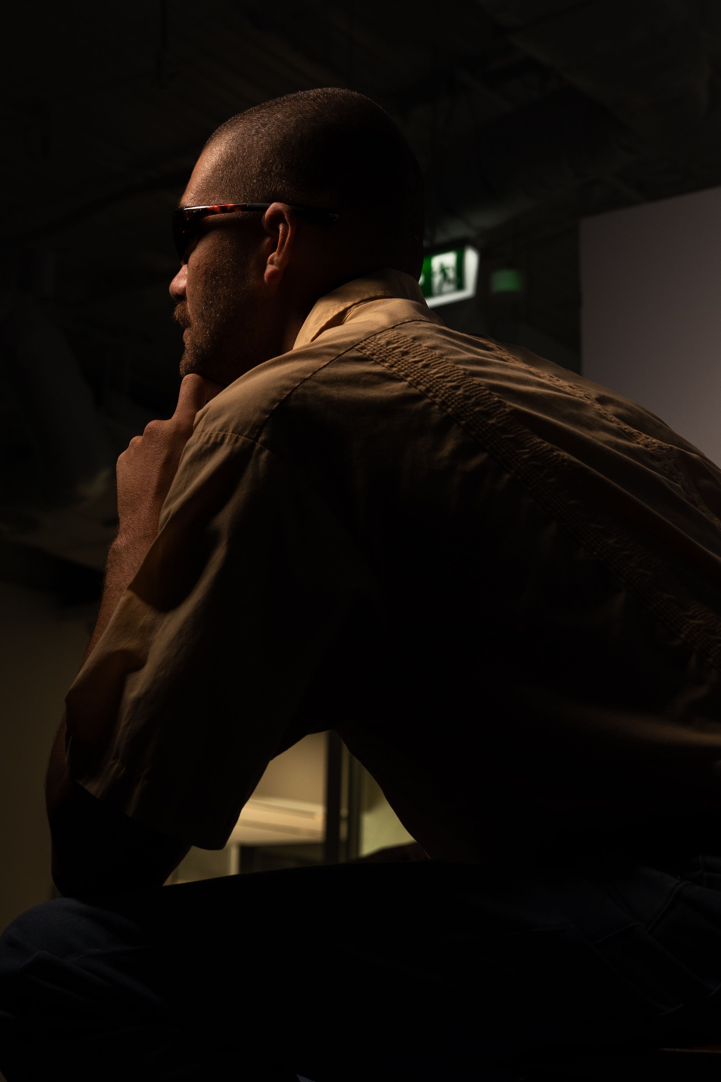Man in olive shirt sitting in contemplative pose in a dimly lit dark environment.