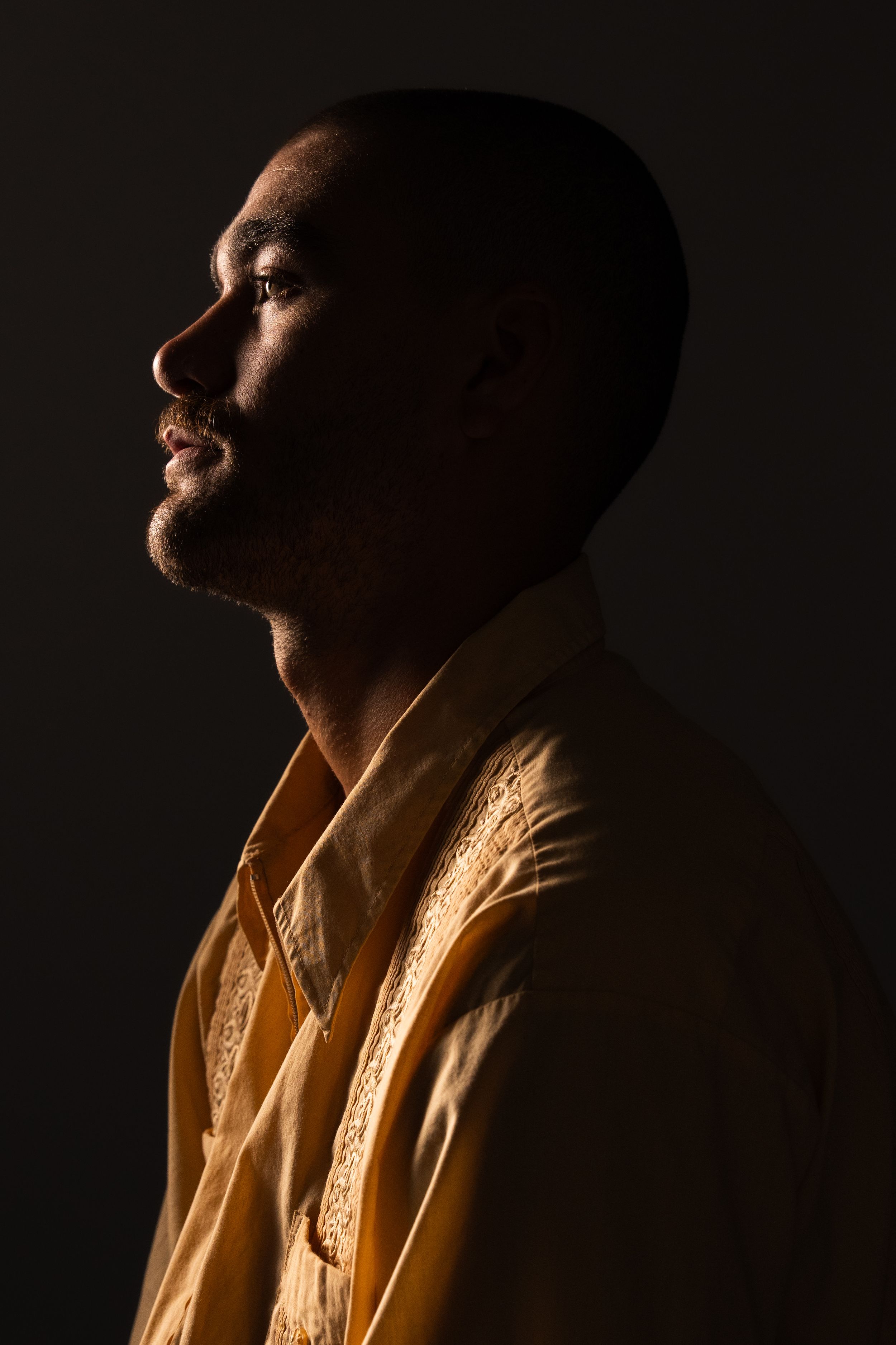 Side profile of a man in a yellow embroidered shirt against a dark dramatic background.