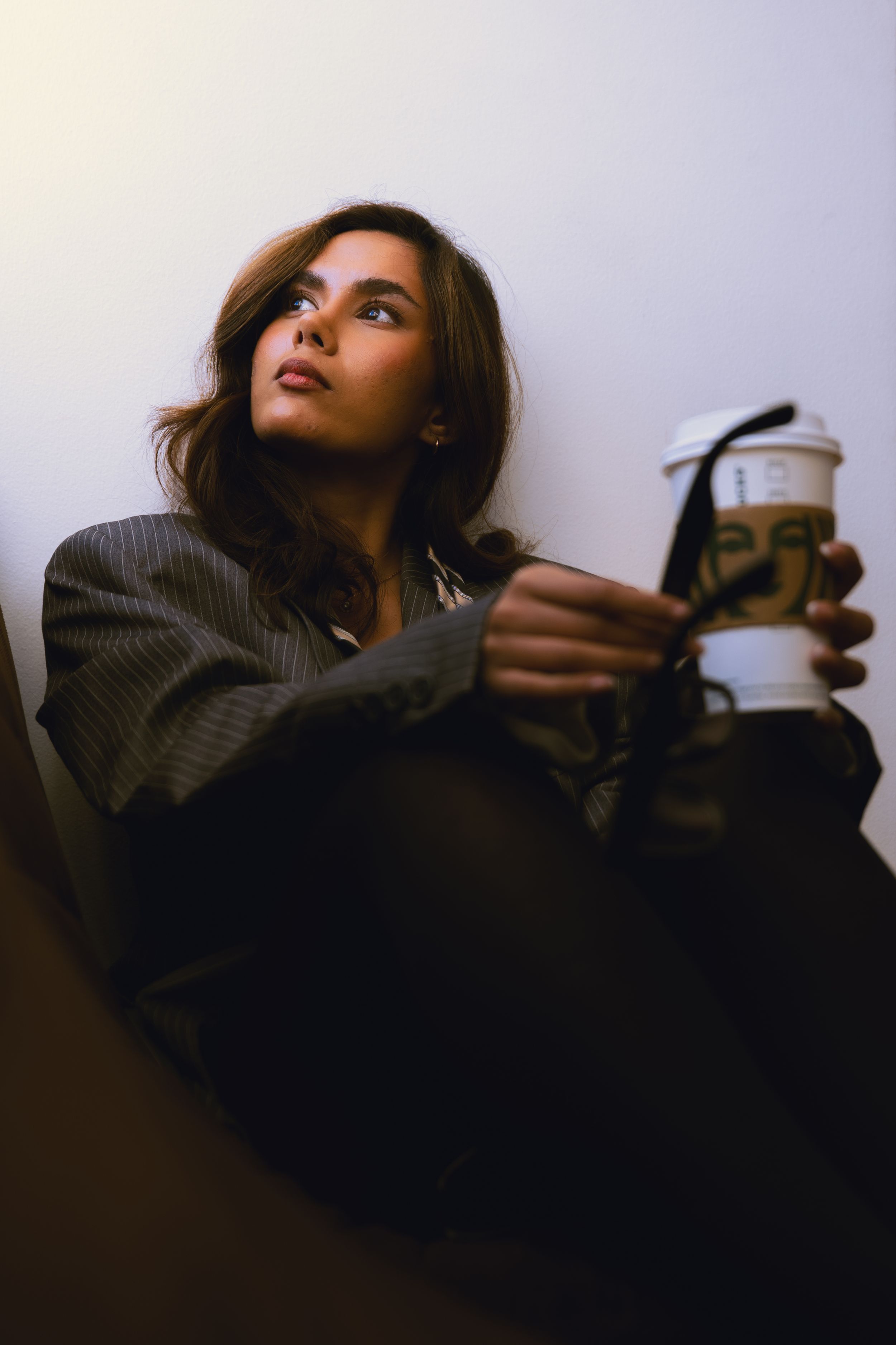 Young woman in grey pinstripe blazer sitting against wall holding takeaway coffee cup looking upward.