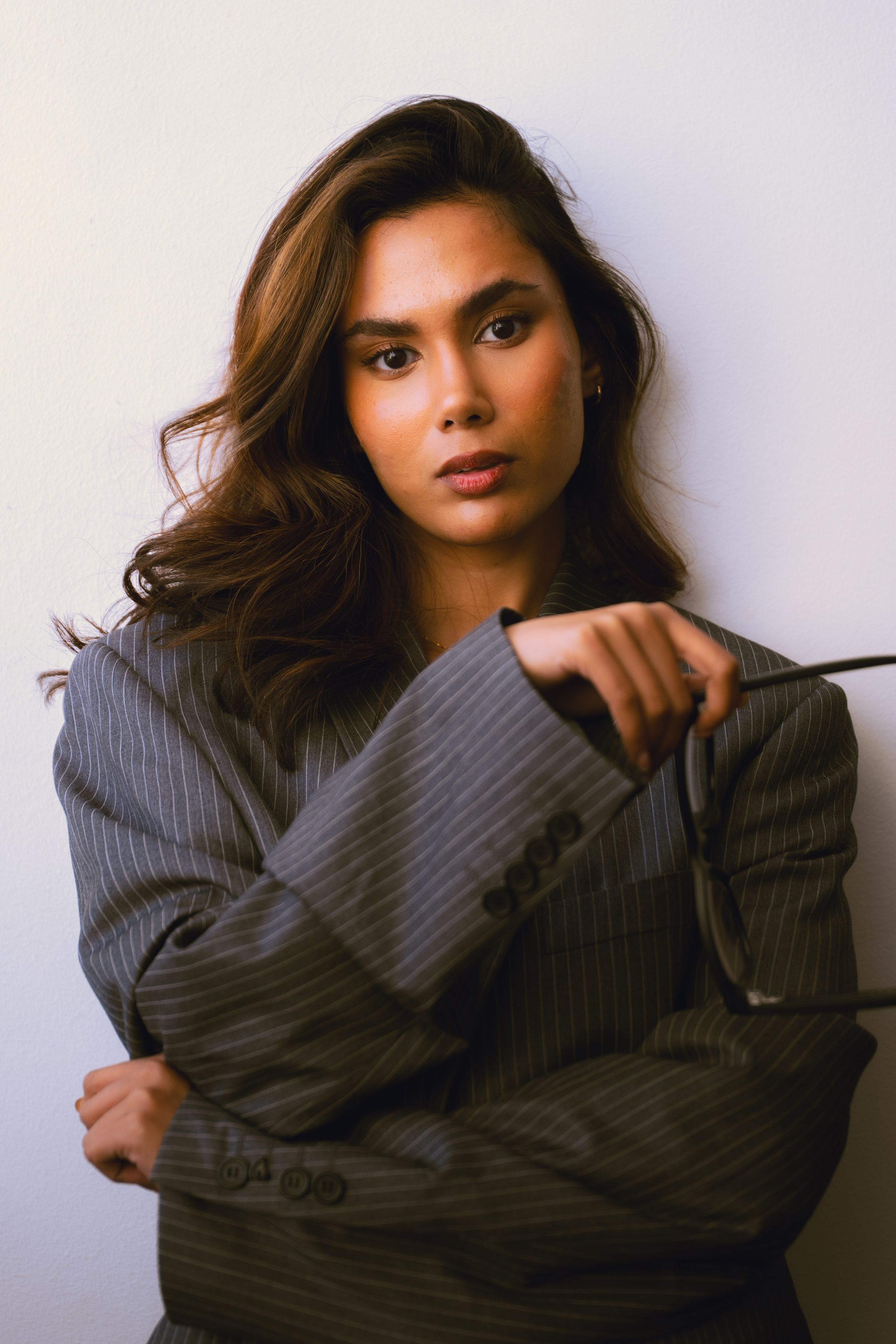 Confident young woman with long brunette hair wearing oversized grey pinstripe blazer posing against white wall.