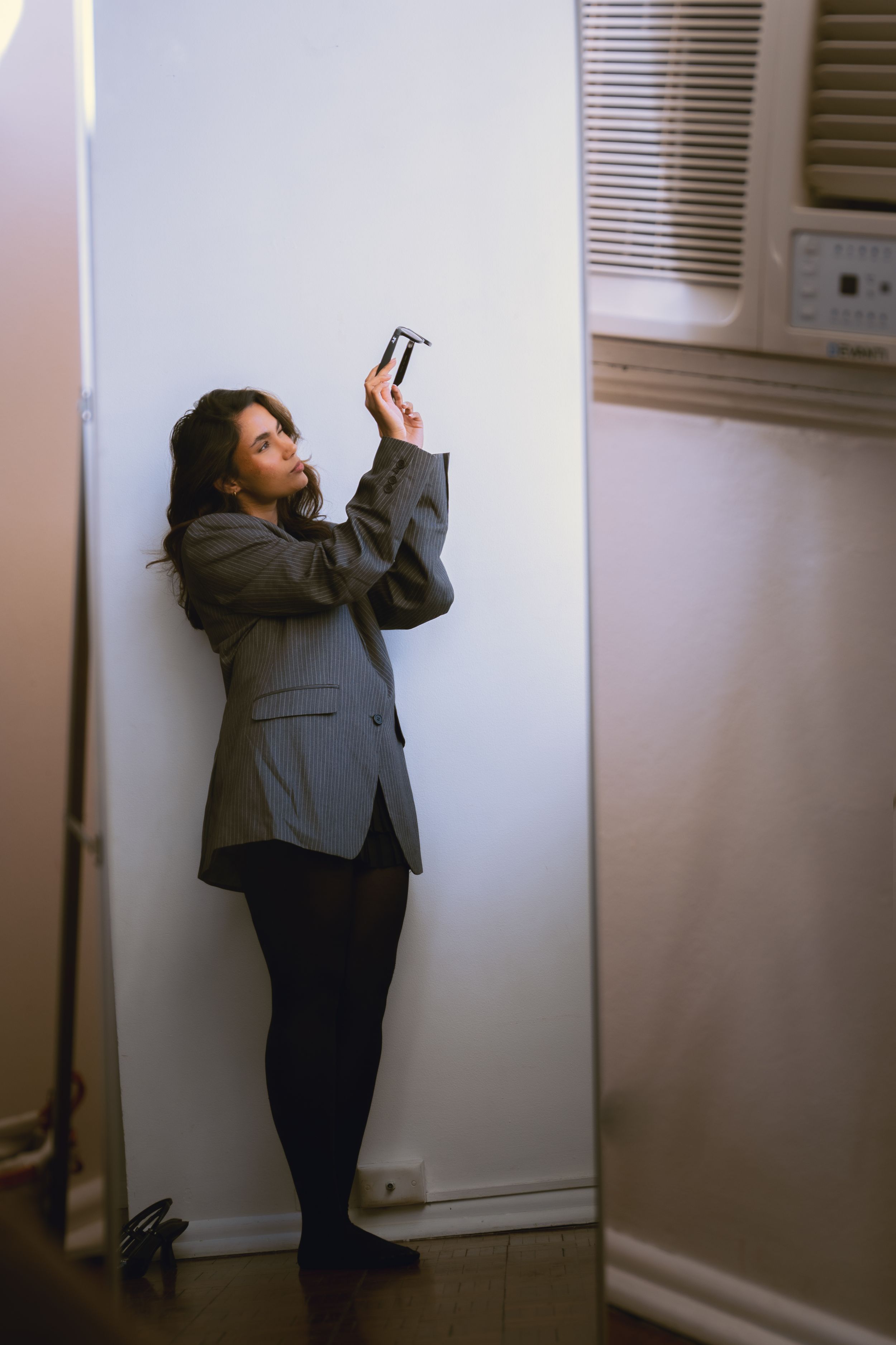 Young brunette woman in grey blazer and black tights taking selfie while leaning against white wall indoors.