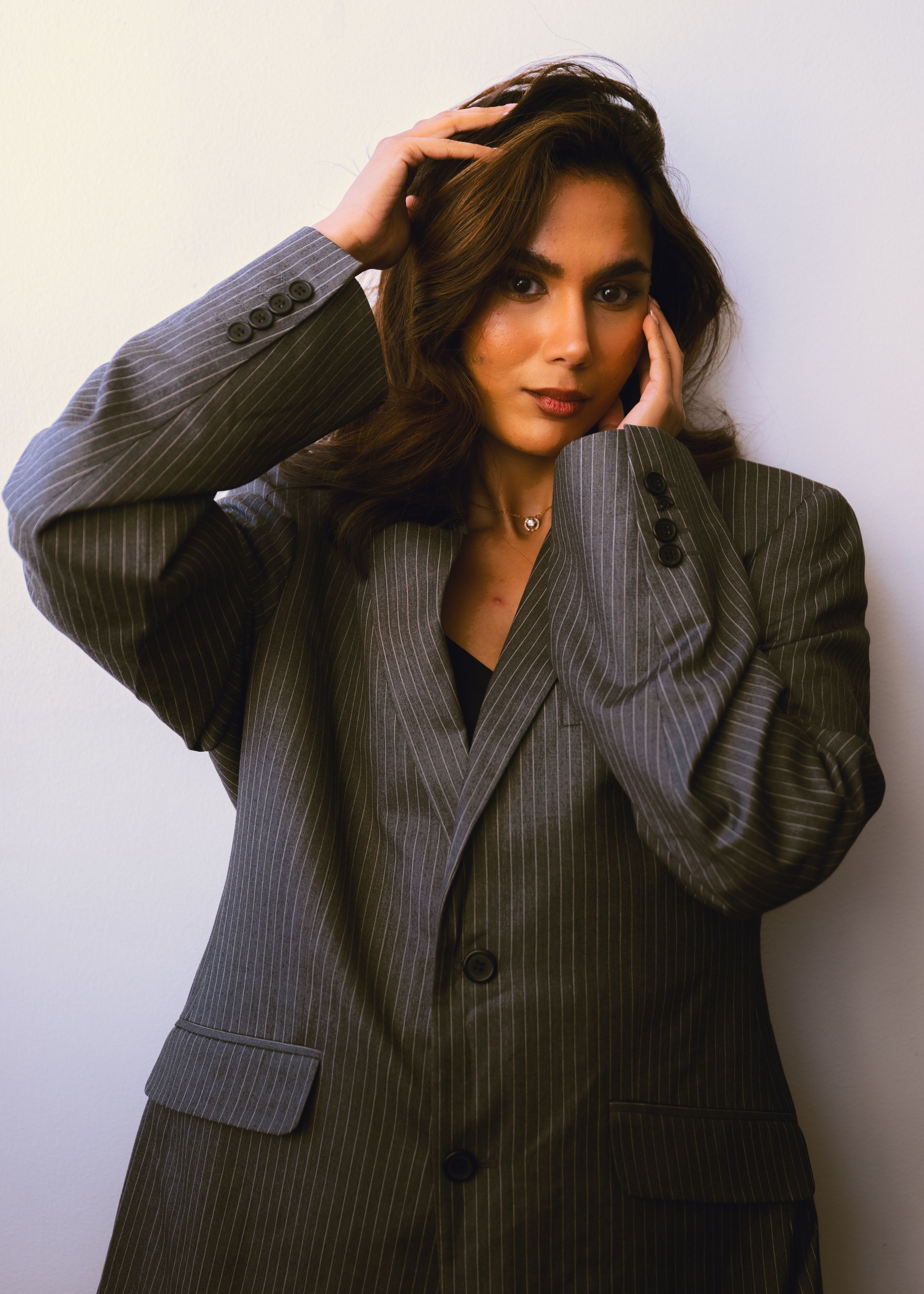 Stylish young woman with long brunette hair in grey pinstripe oversized blazer posing with hands in hair.